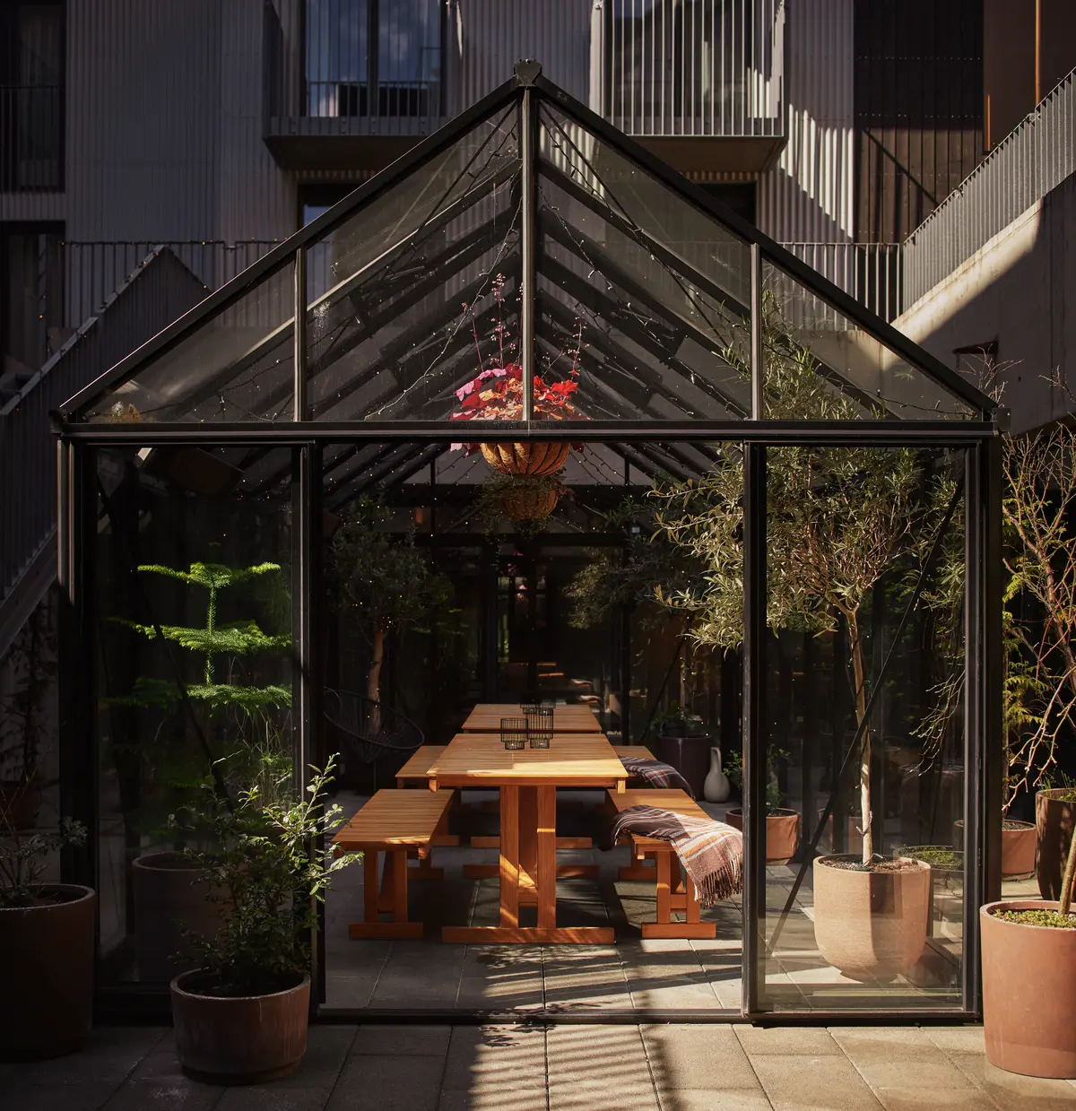 Sunlit glass garden house with long wooden table and potted plants.
