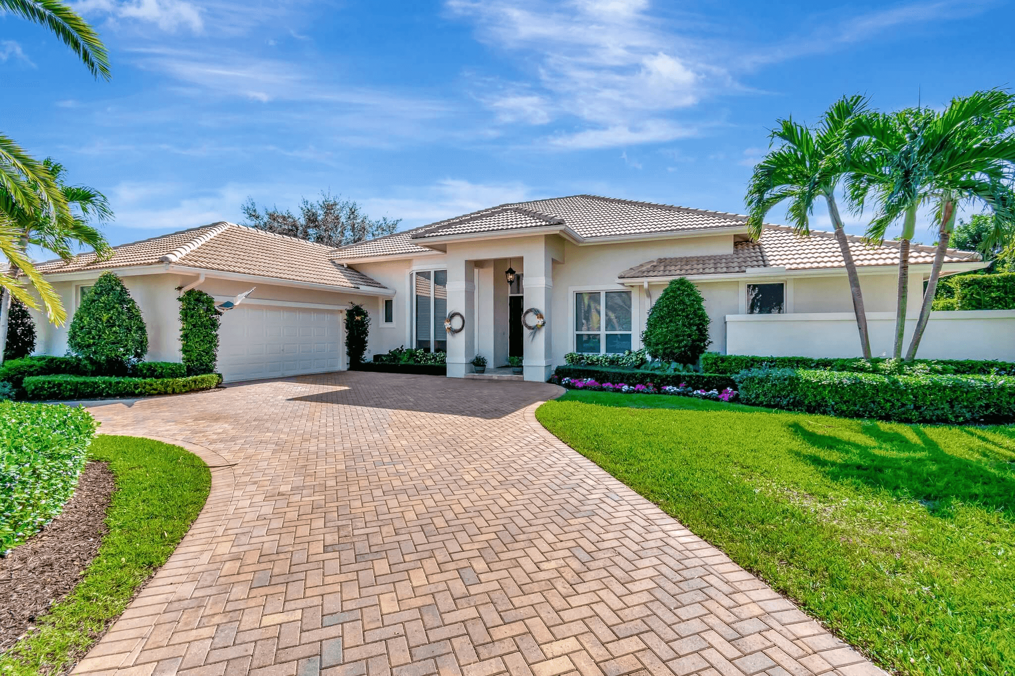 Single-story Mediterranean-style home with clay tile roof, cream stucco exterior, and curved paver driveway. Professionally landscaped front yard features manicured hedges, tropical palm trees, colorful flower beds, and lush green lawn. Two columns frame the elegant entryway under bright blue Florida sky.