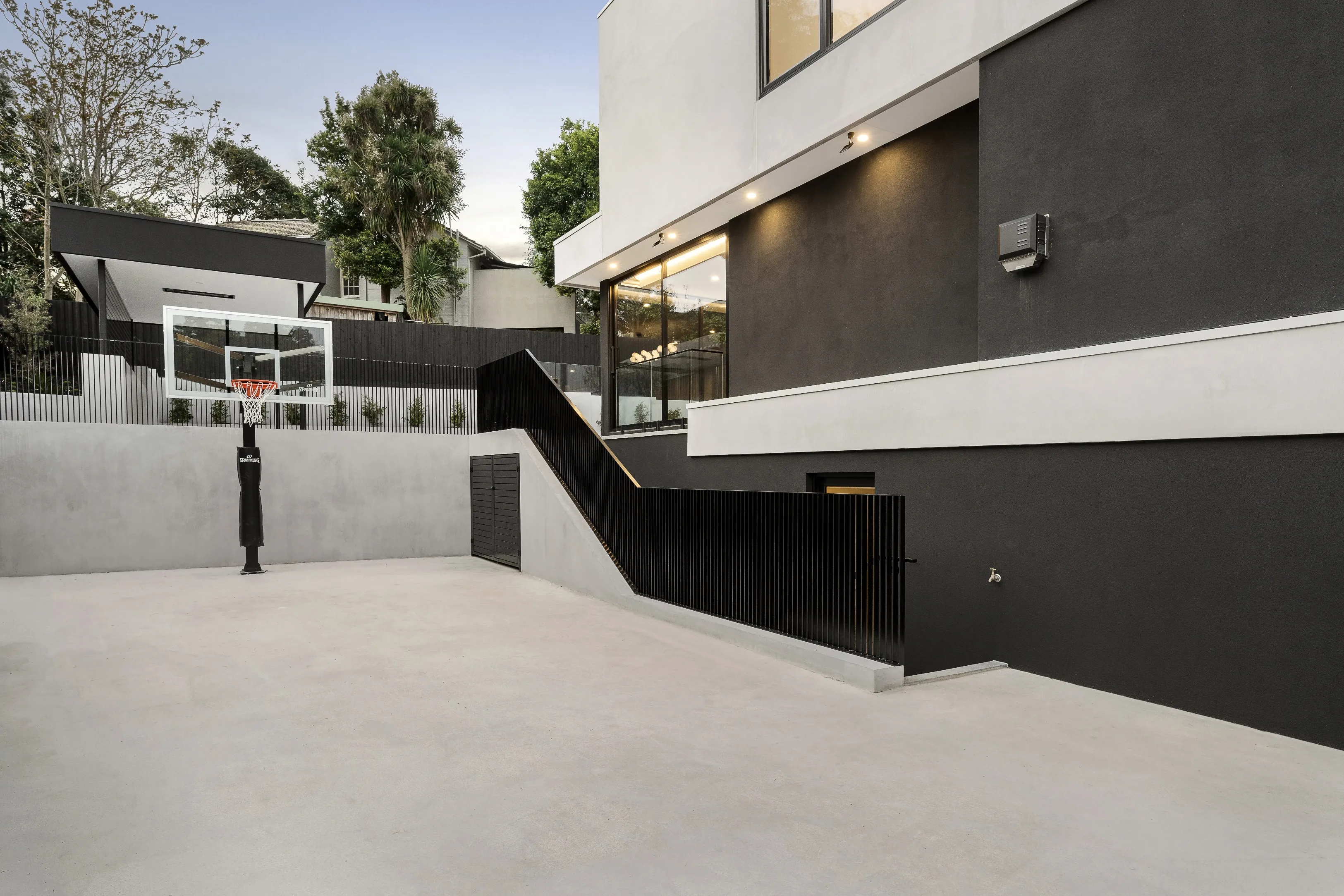 Rear of Ivanhoe East Residence at dusk with a rendered half-basketball court, Spalding hoop, charcoal render and a ramp with black balustrade
