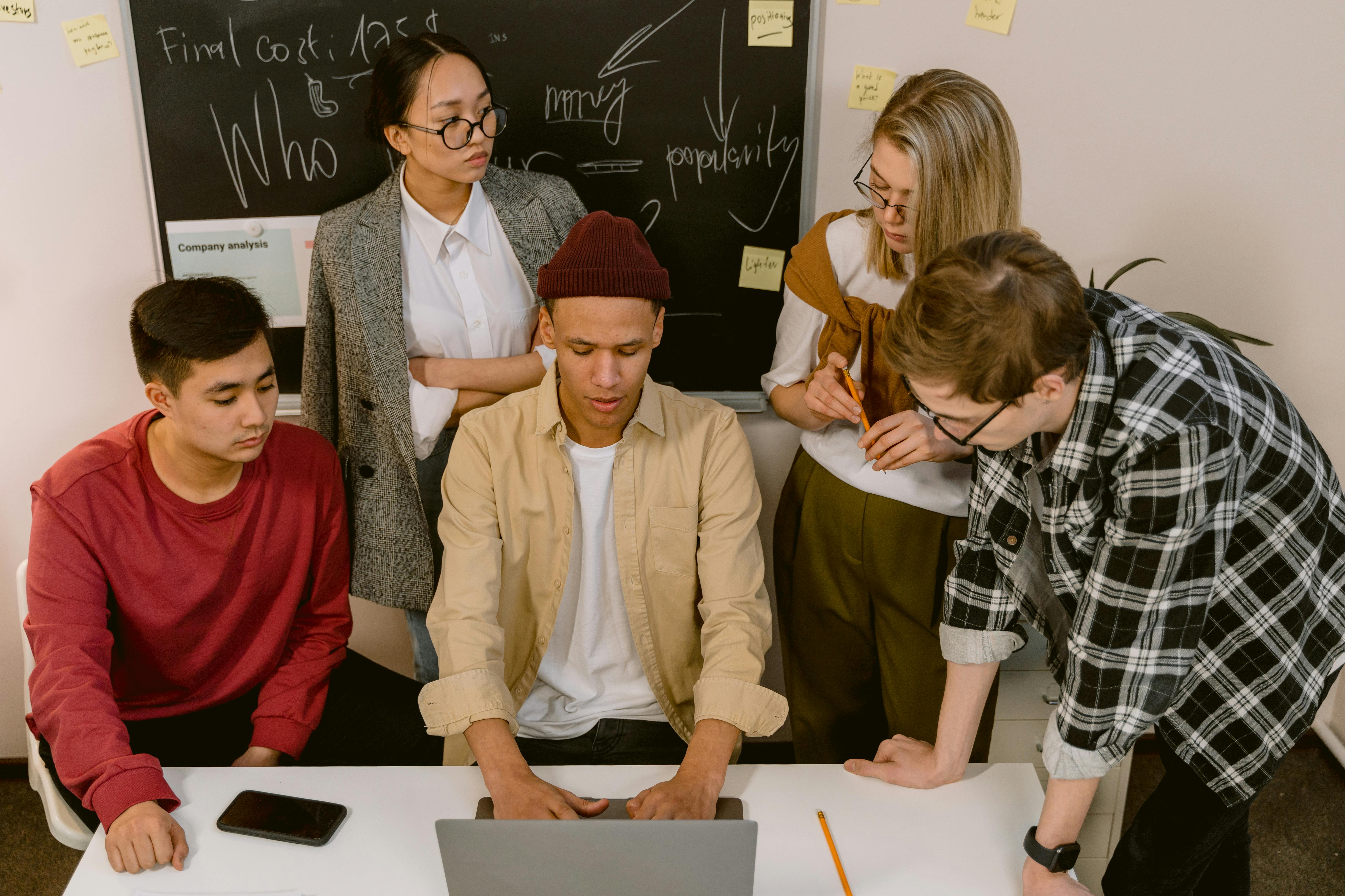 Gruppo di giovani al lavoro davanti a un computer con lavagna piena di appunti alle spalle, rappresentazione della pressione del confronto sociale e del tentativo di “capire cosa fare” osservando le scelte degli altri.