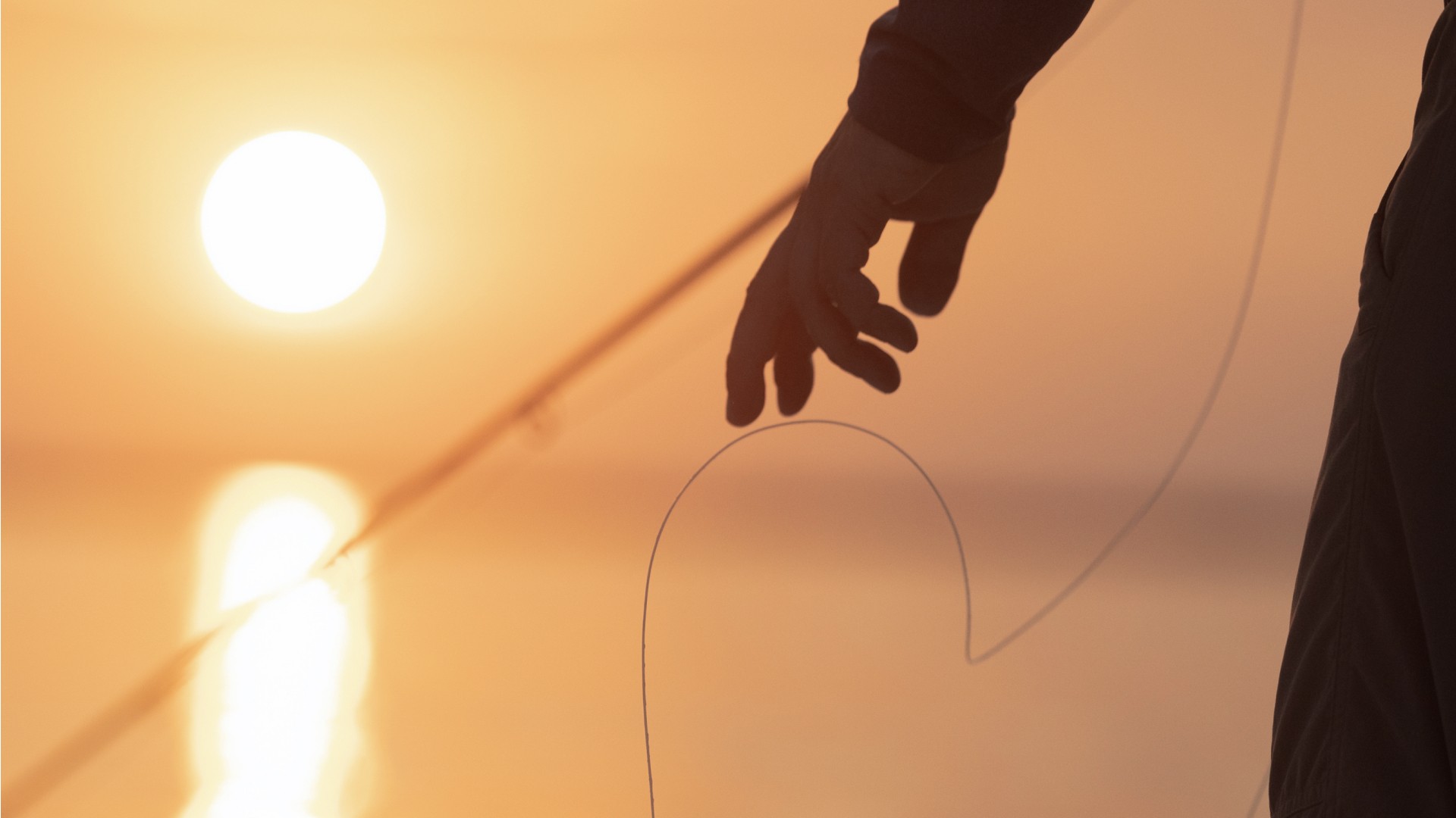 Detail of a hand and a fly fishing rod with the sunset and glowy yellow light in the background