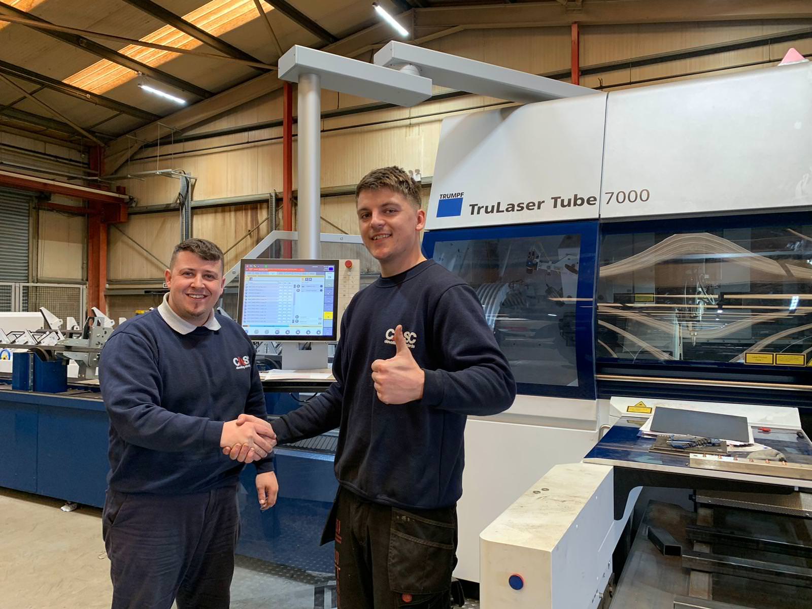 Two people shaking hands inside a workshop in front of an industrial tube laser cutting machine.