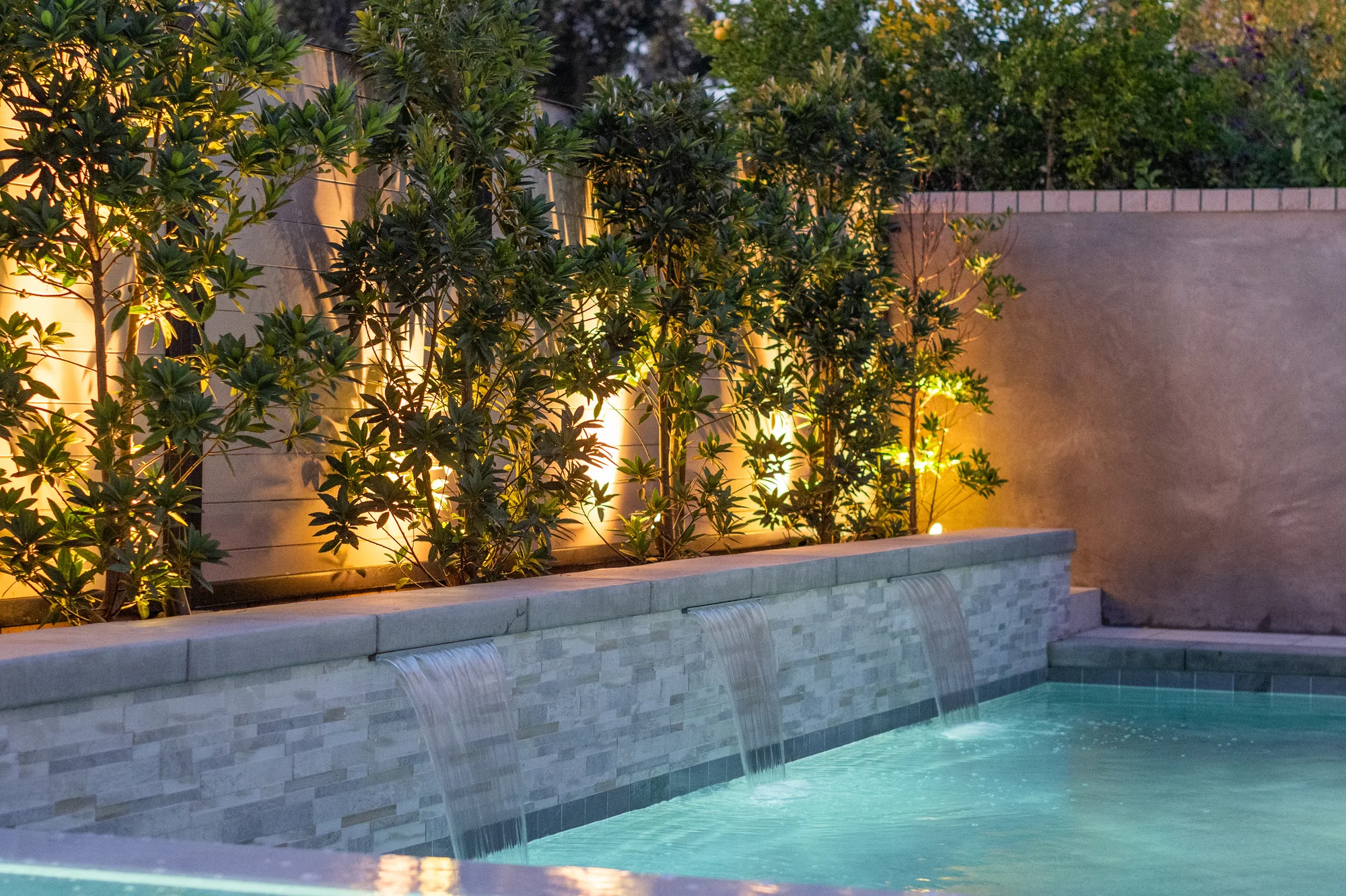 Waterfall spouts in a stone-faced pool wall below illuminated shrubs.