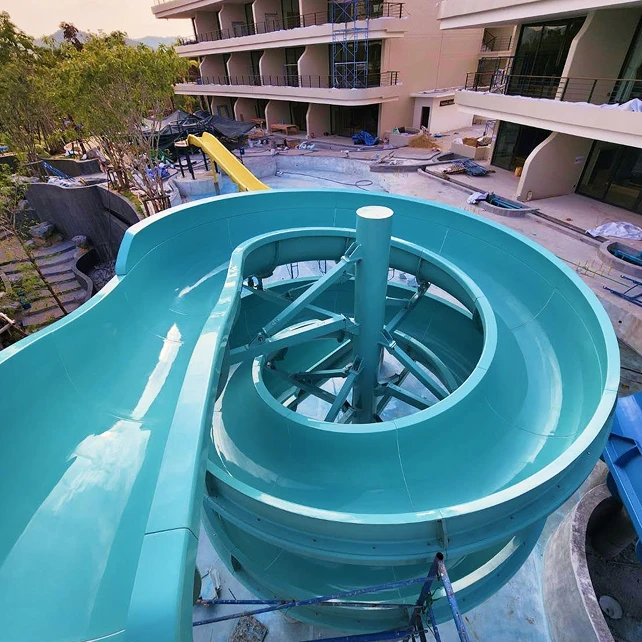 Một máng trượt nước xoắn ốc màu xanh lục nhạt nằm phía trên khu vực hồ bơi chưa hoàn thiện của khu nghỉ dưỡng, được bao quanh bởi các tòa nhà màu be và cây cối. Cảnh tượng này mang đến cảm giác mong đợi và sự nhộn nhịp.