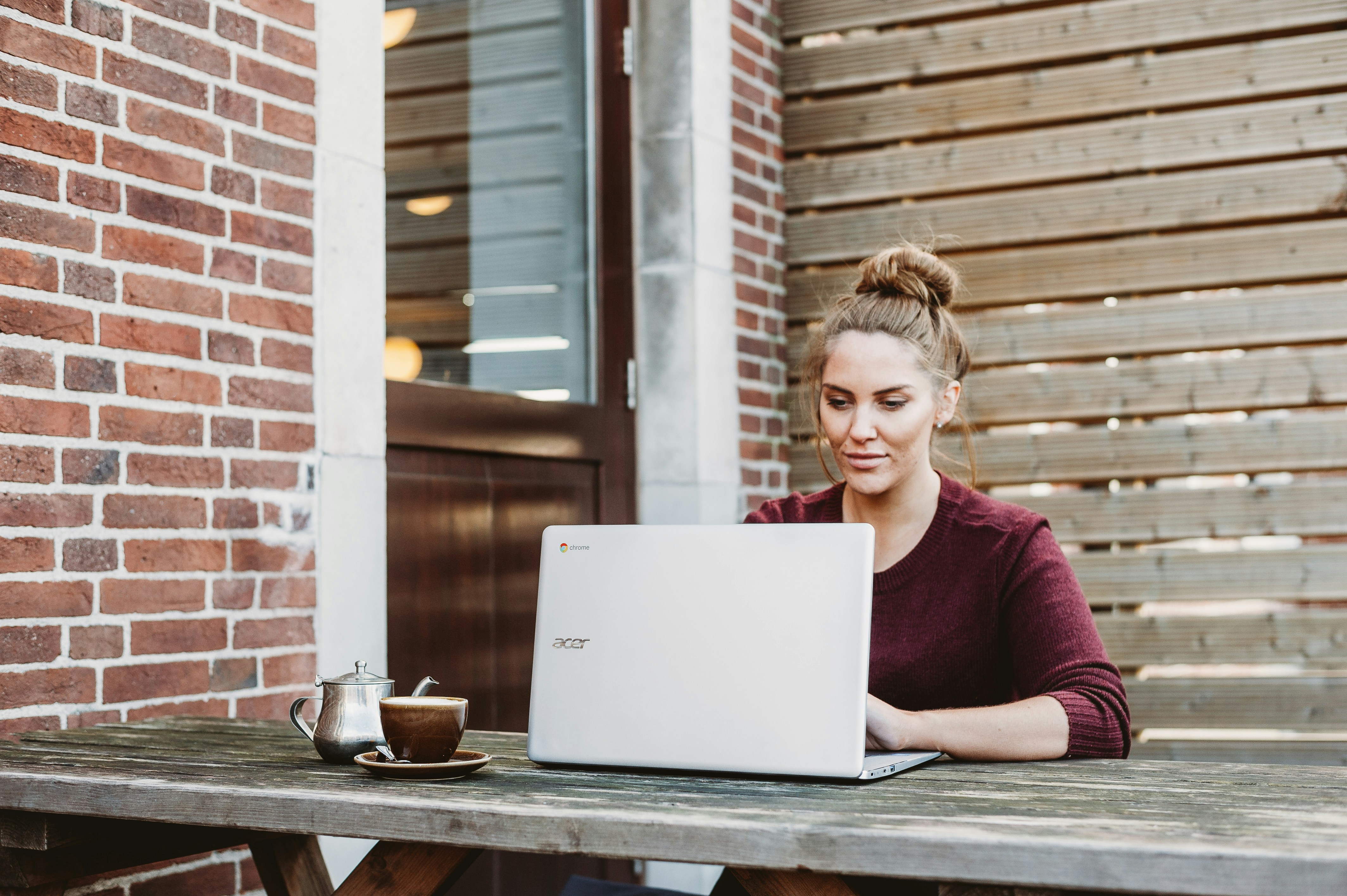 A young woman sits at a table outside with a laptop open infront of her. There is a mug by the laptop. 