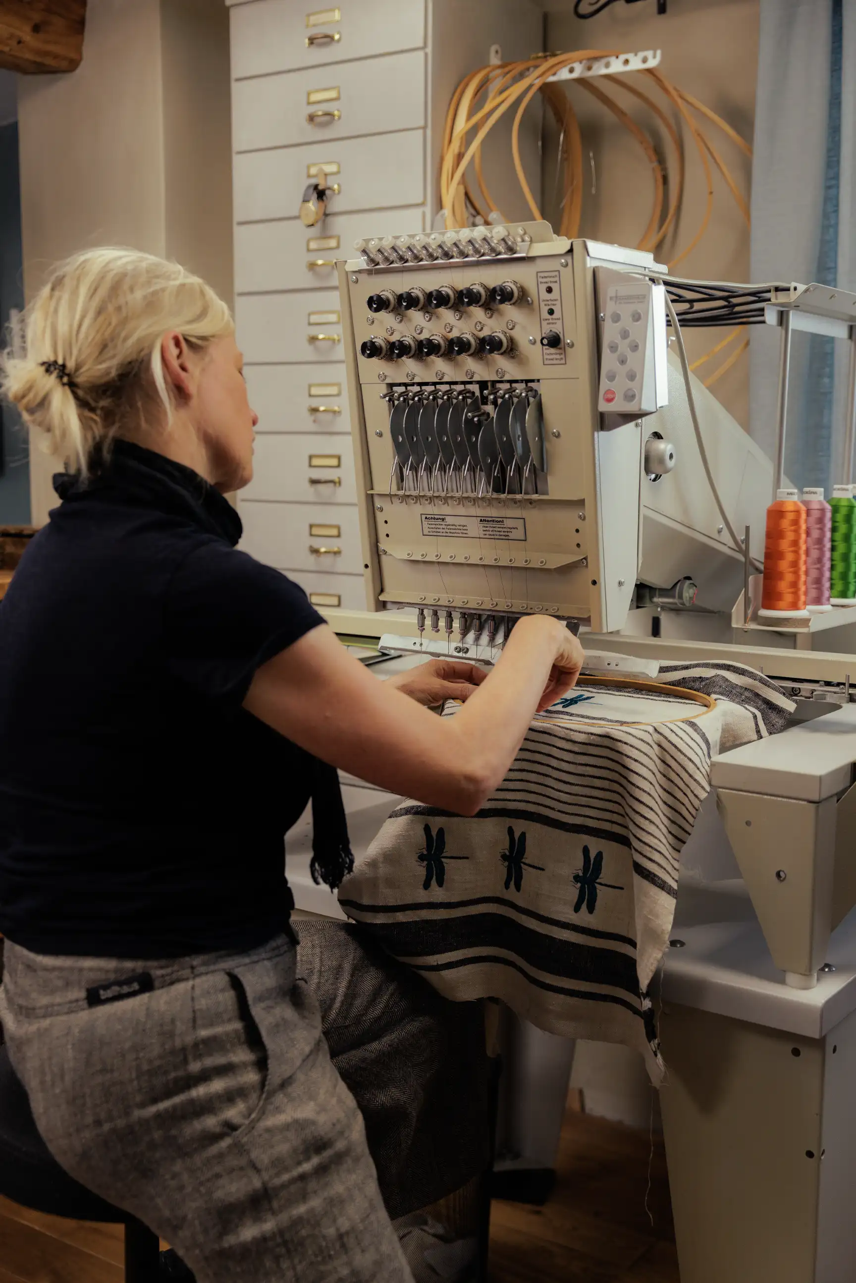 Eine Frau arbeitet an einer Stickmaschine im Atelier Wildwechsel Leipzig, während sie ein gestreiftes Leinentuch mit Libellenmotiv bestickt.