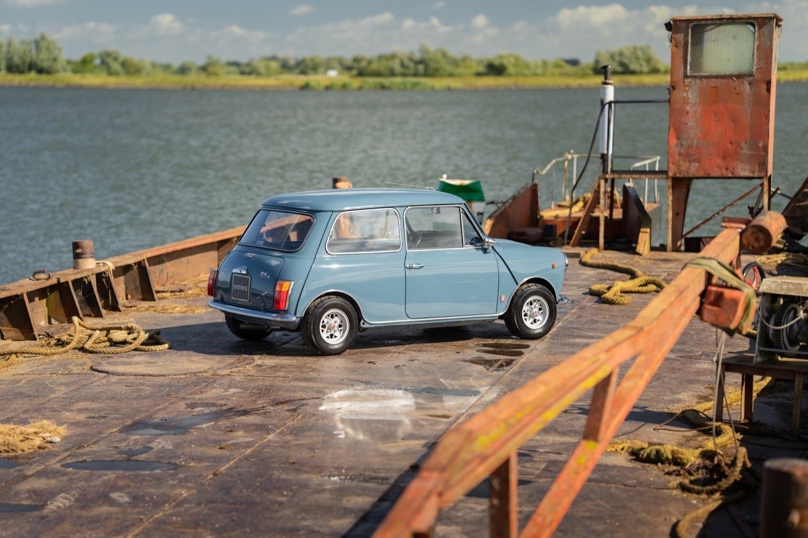 A vintage blue car parked on a wooden dock by the water, with a ferry and scenic landscape in the background.