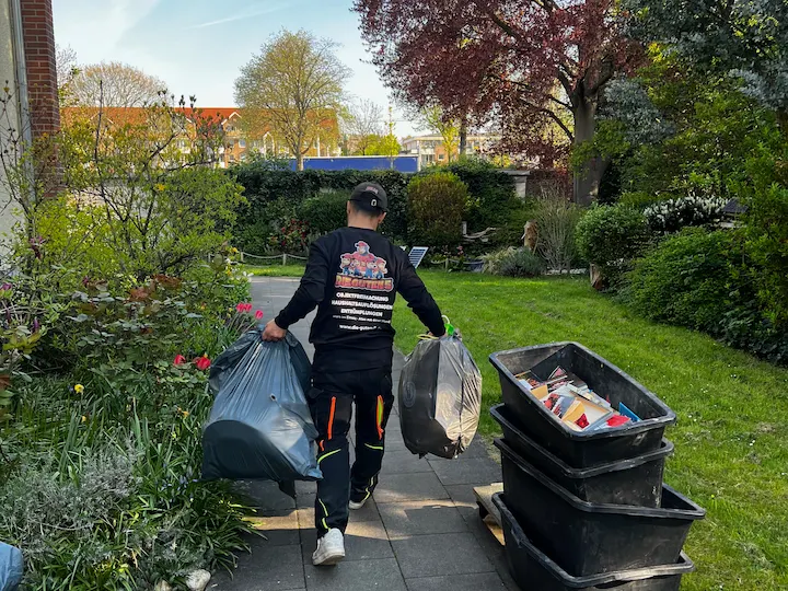 Mitarbeiter trägt Müllsäcke durch einen Vorgarten neben Wannen mit Sperrmüll