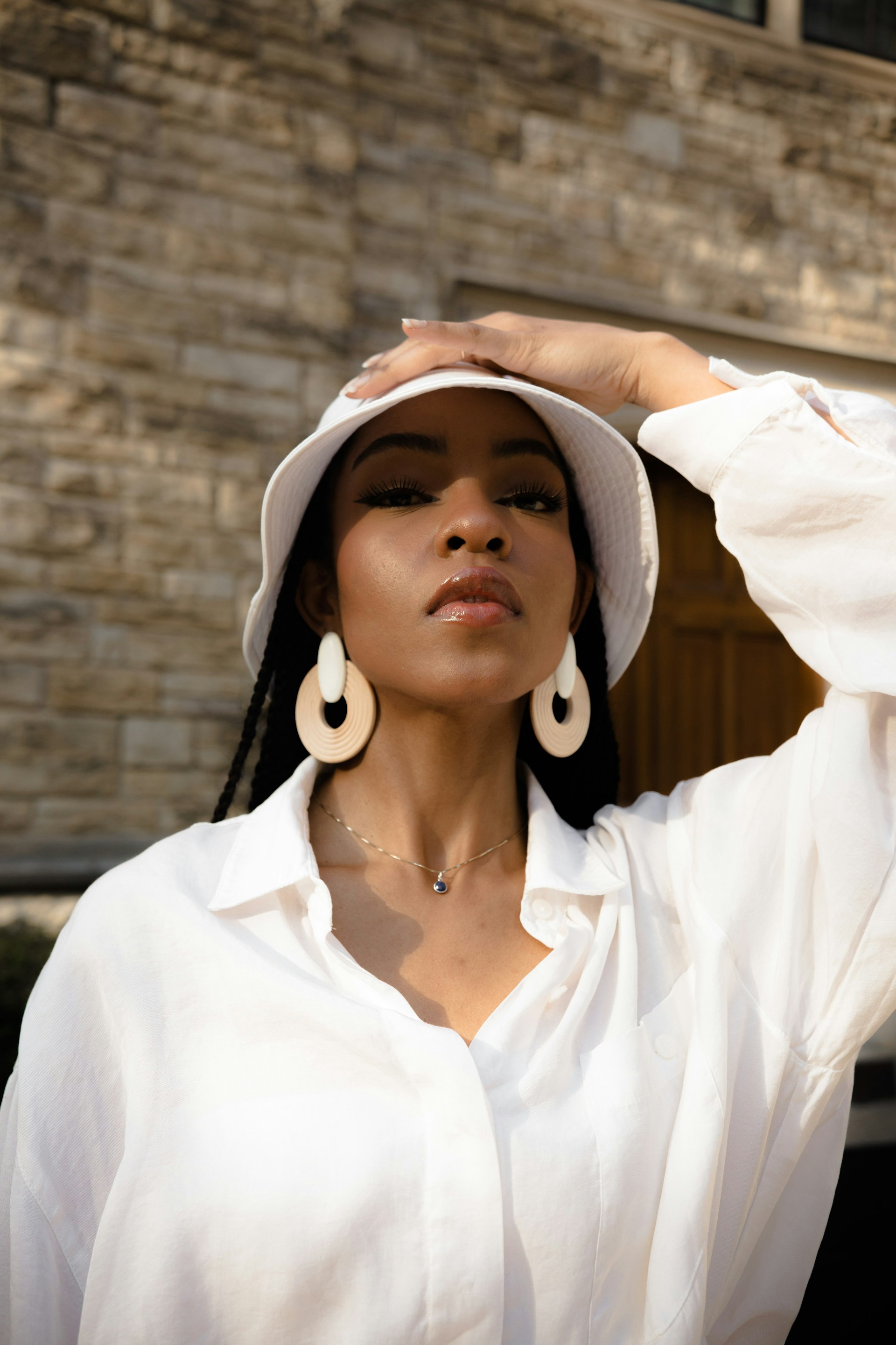 A woman in a white bucket hat and shirt wearing large cream-colored drop earrings and a dainty pendant necklace.