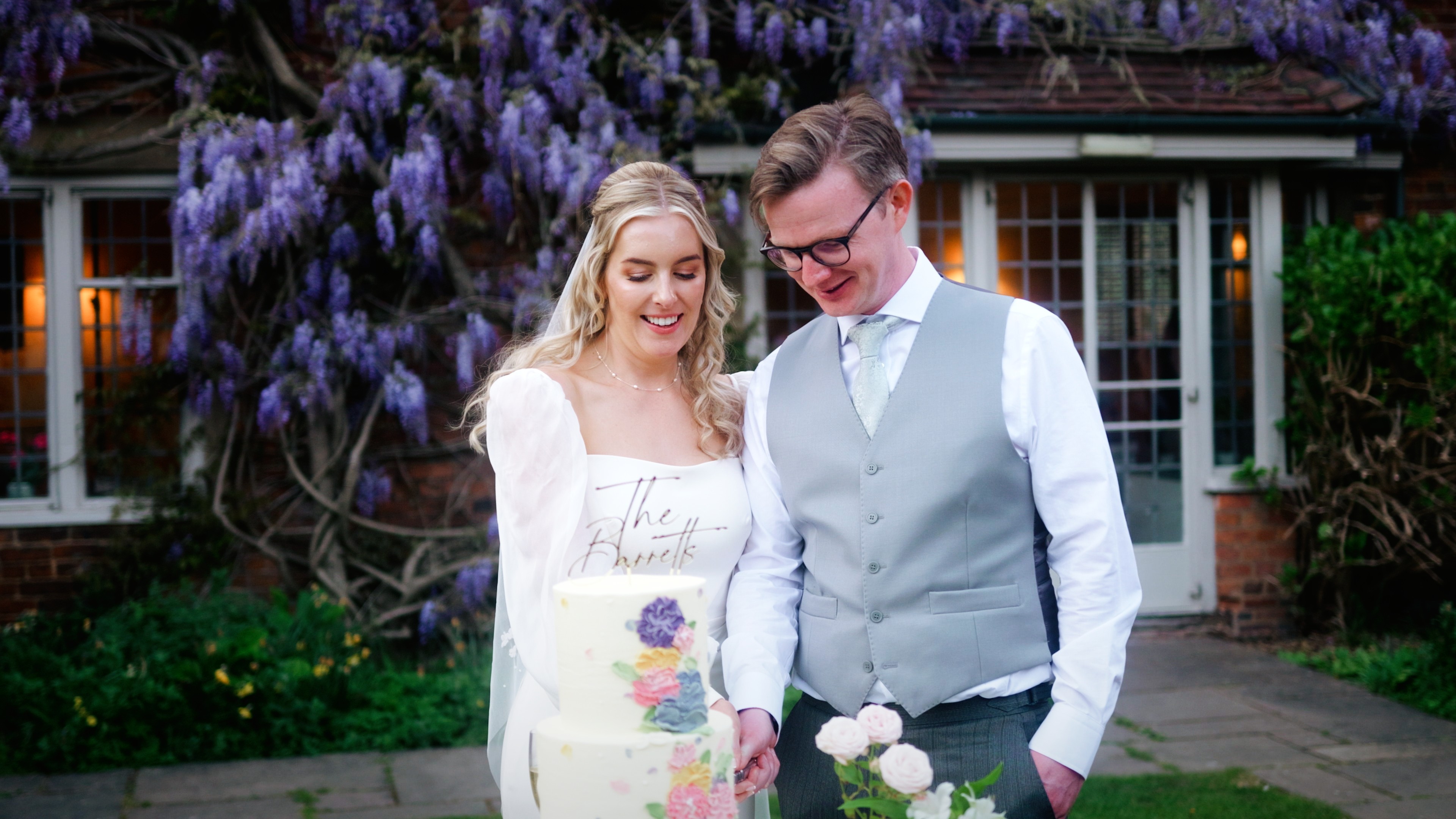 Bride and groom cutting their wedding cake outside at Doddington Hall in Lincolnshire, with wisteria and the house behind them