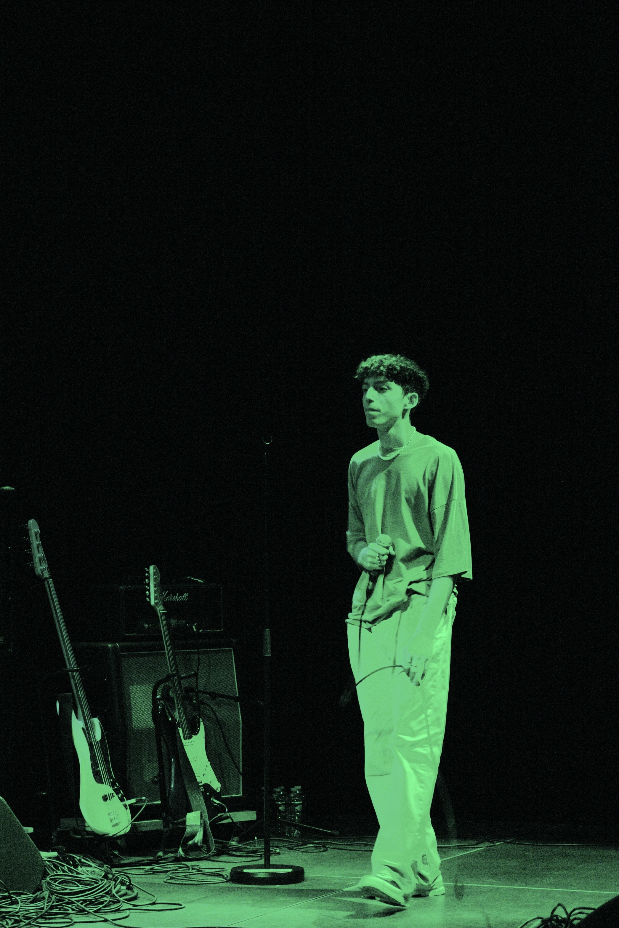 A performer stands in green lighting, with guitars and musical equipment in the shadows behind them.