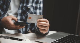 A man at a desk holds a credit card in one hand and a cell phone in the other. On the desk is a laptop and pen and paper.