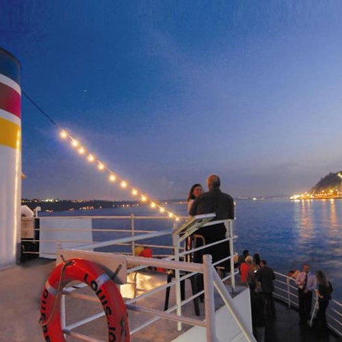 Mọi người đang tụ tập trên boong tàu vào lúc chạng vạng, với đèn dây lấp lánh trên đầu và bờ biển cùng mặt nước hiển thị phía sau.