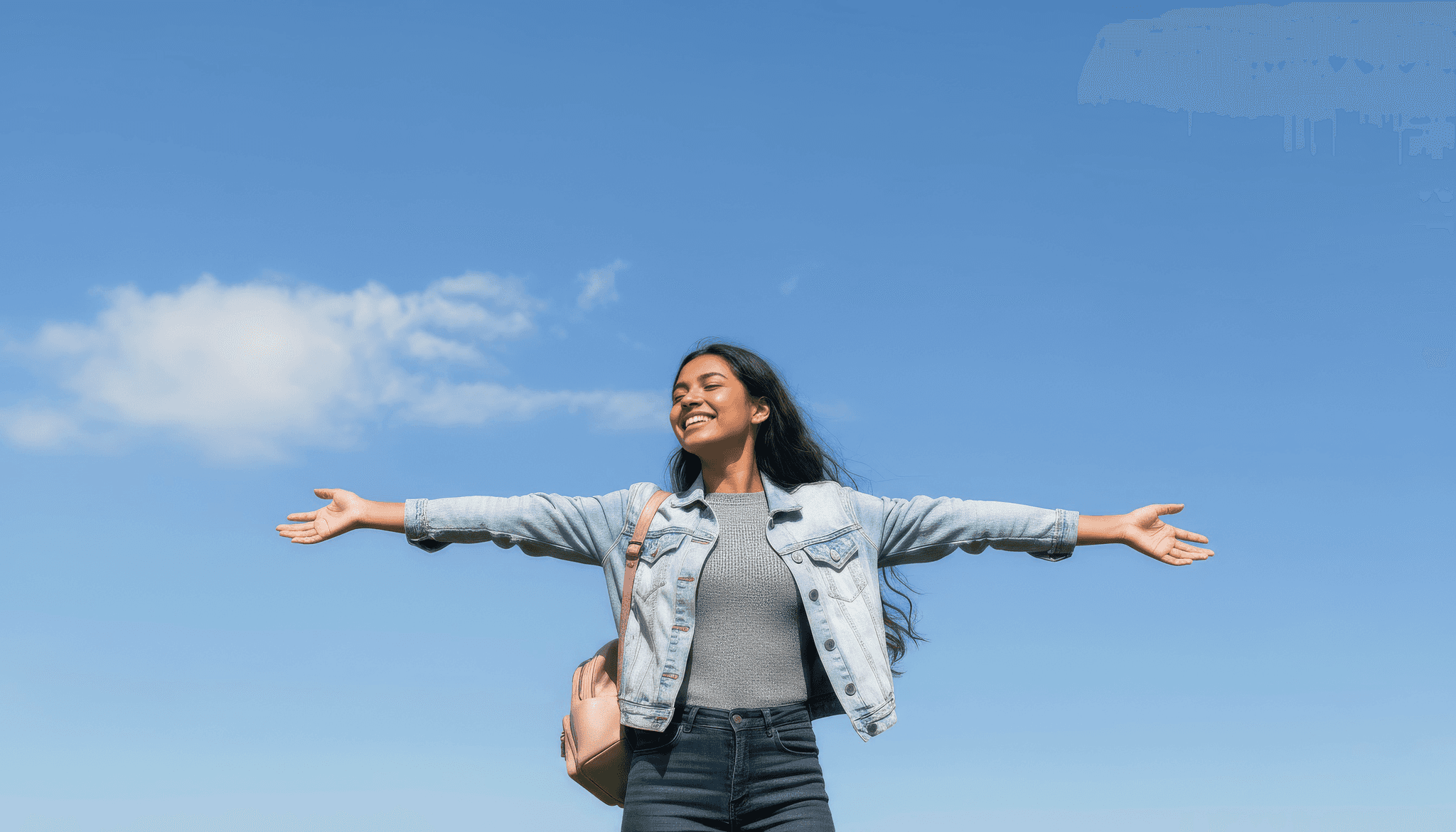 A female student stands in the open air, arms outstretched, with clear blue skies and a few clouds overhead.