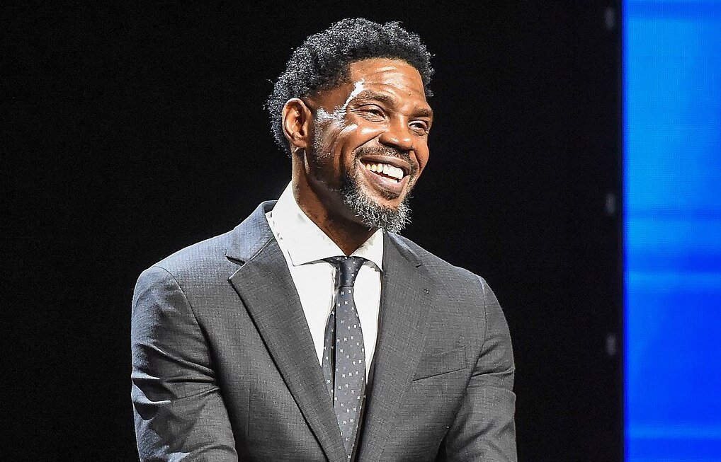 Udonis Haslem with short curly hair and a beard, wearing a dark suit, white shirt, and polka dot tie, smiles warmly against a dark background with a blue panel on the right.