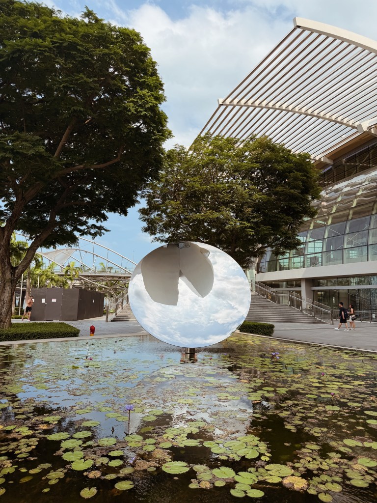 A modern sculpture in a reflective, circular shape sits in a pond filled with lily pads, surrounded by lush trees and contemporary architecture.