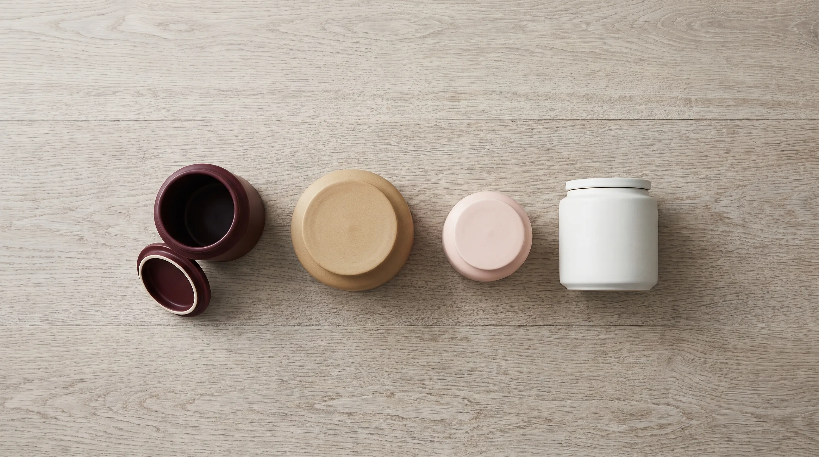 Four matte stoneware jars in a row on a pale oak table seen from directly above, three closed and the burgundy one open with its lid resting beside it