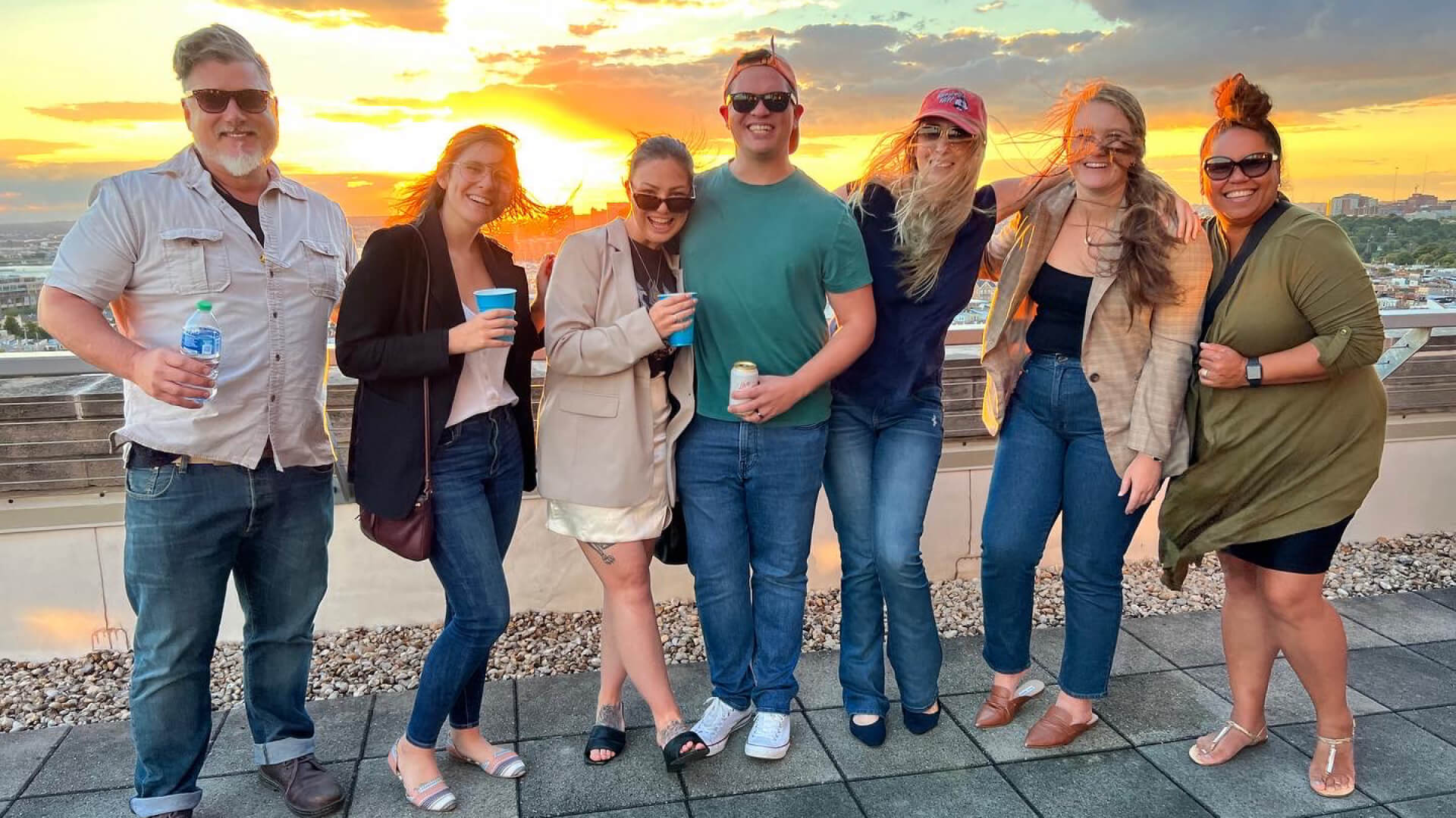 Seven people stand on a rooftop at sunset, smiling at the camera; some hold drinks, and the city skyline is visible in the background.