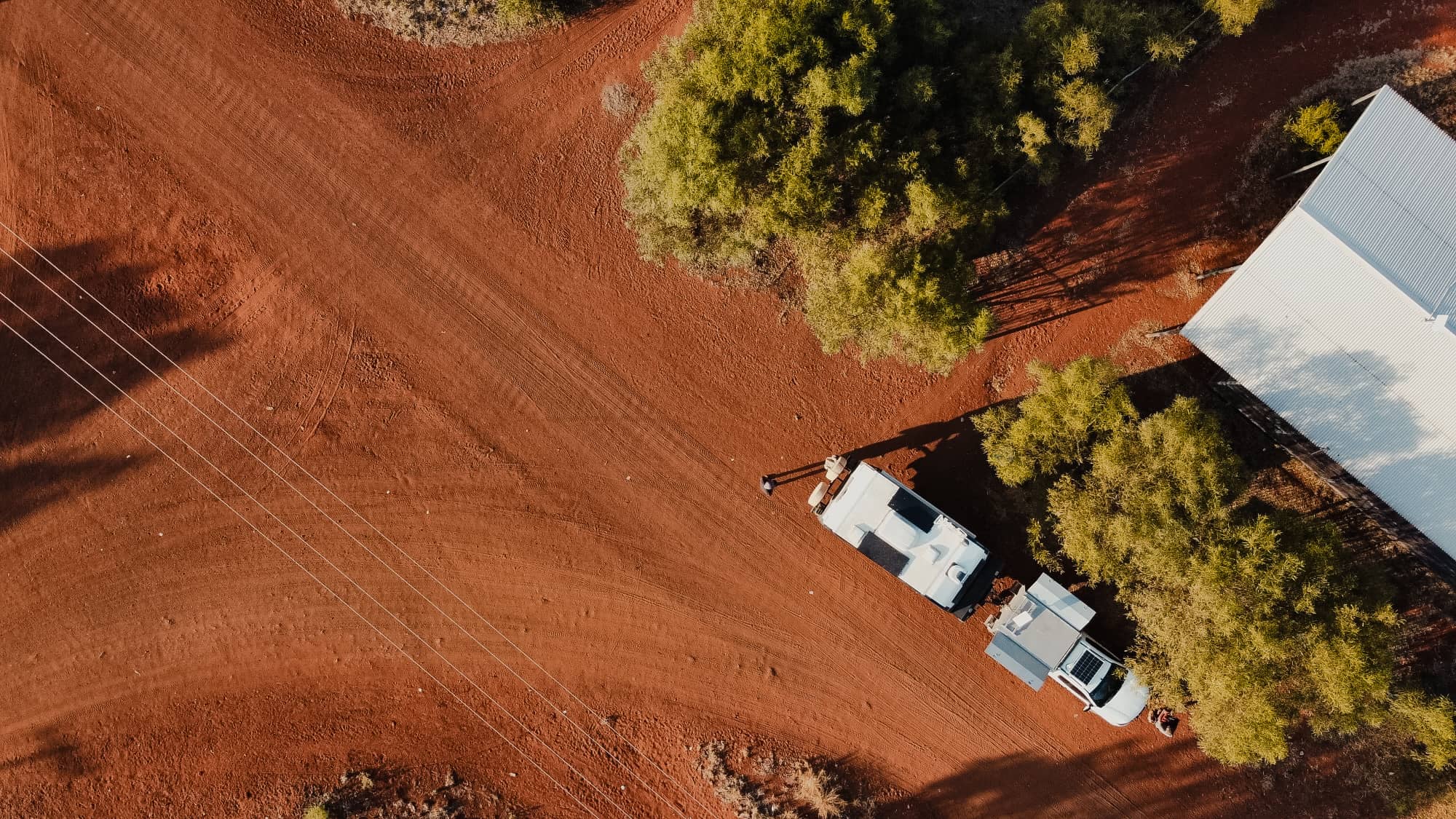 Aerial view of a remote Australian outback setting with red dirt, sparse trees, a white building, and a caravan and SUV.