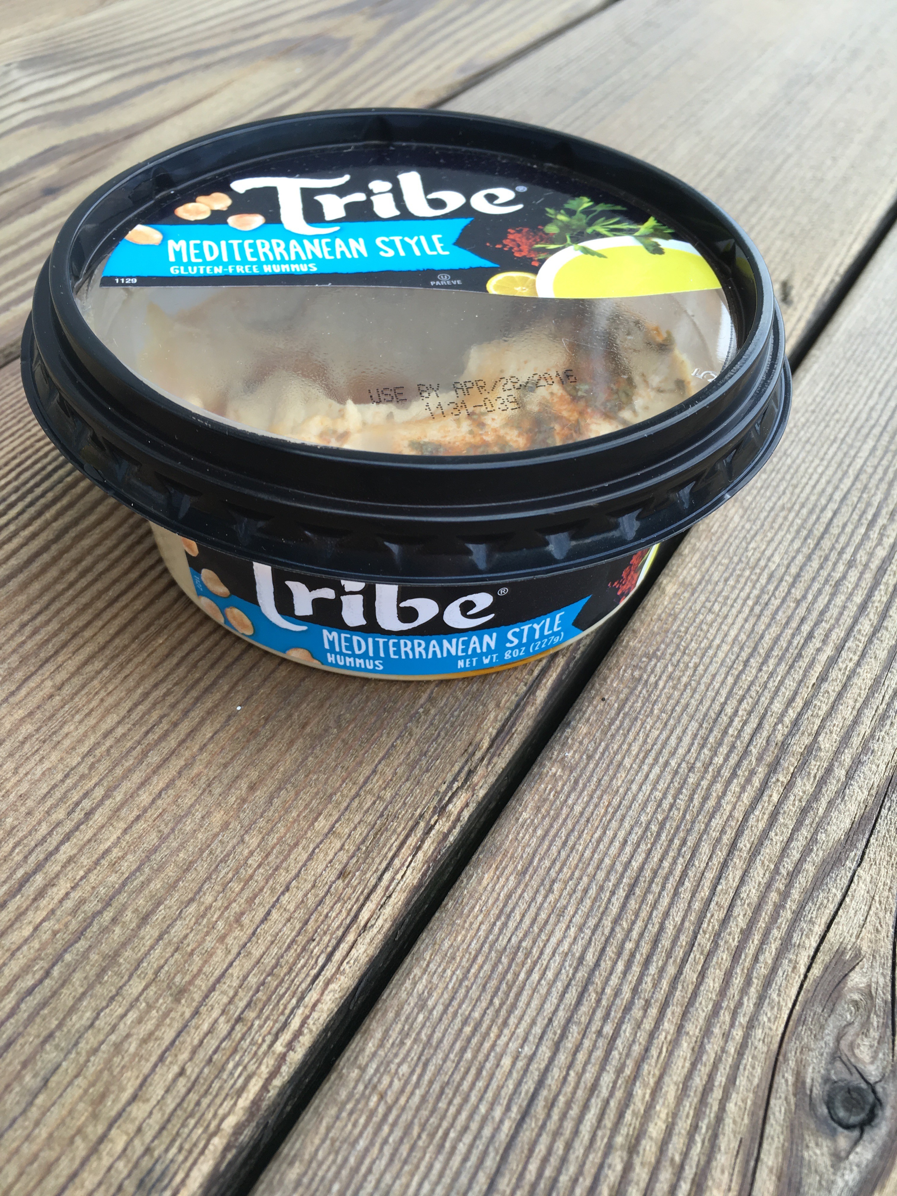 A container of Tribe Mediterranean Style hummus with a clear lid revealing a creamy, spiced dip, placed on a rustic wooden surface.