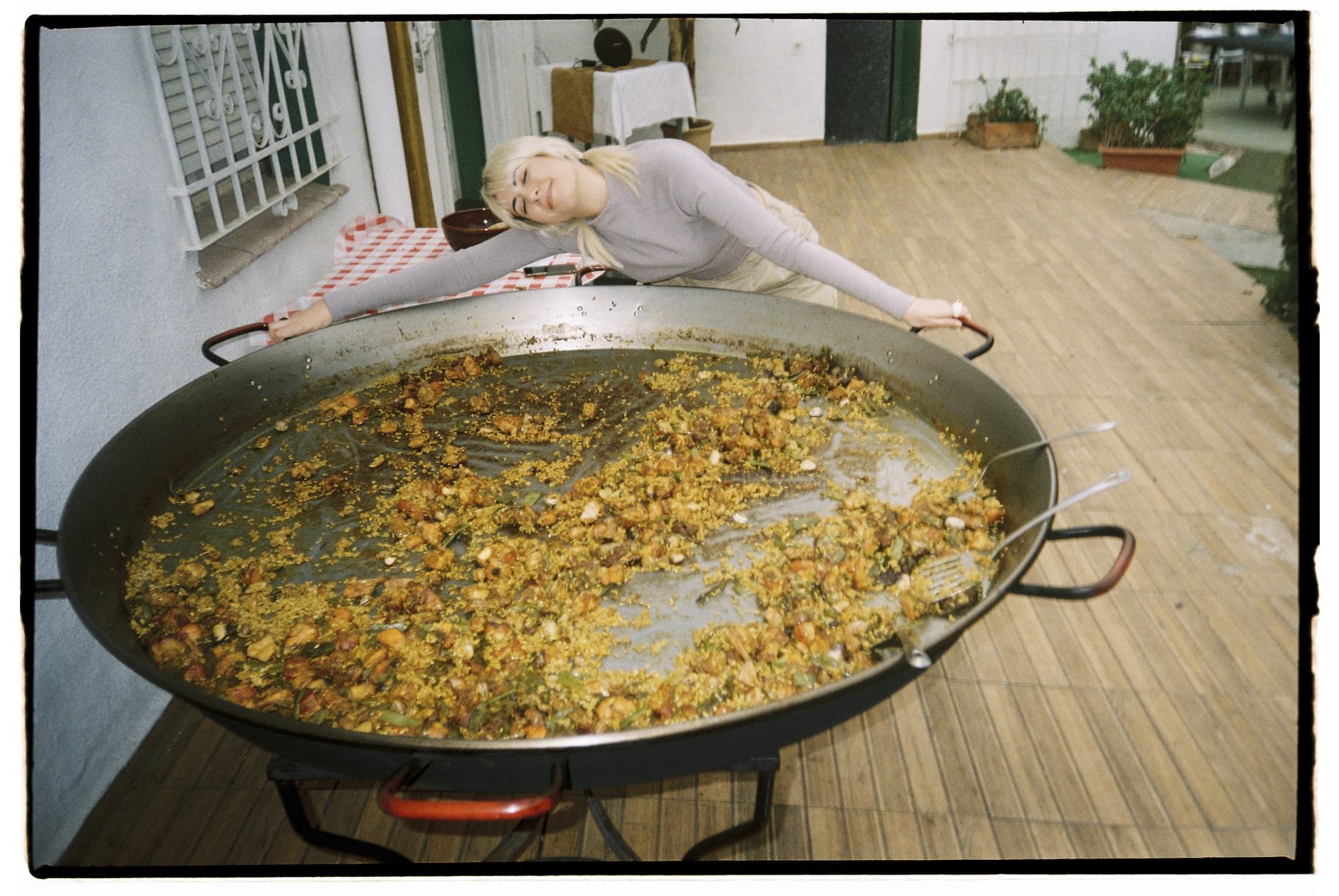 A woman with blonde hair leaning over a large paella pan with remaining rice and vegetables at an outdoor patio.