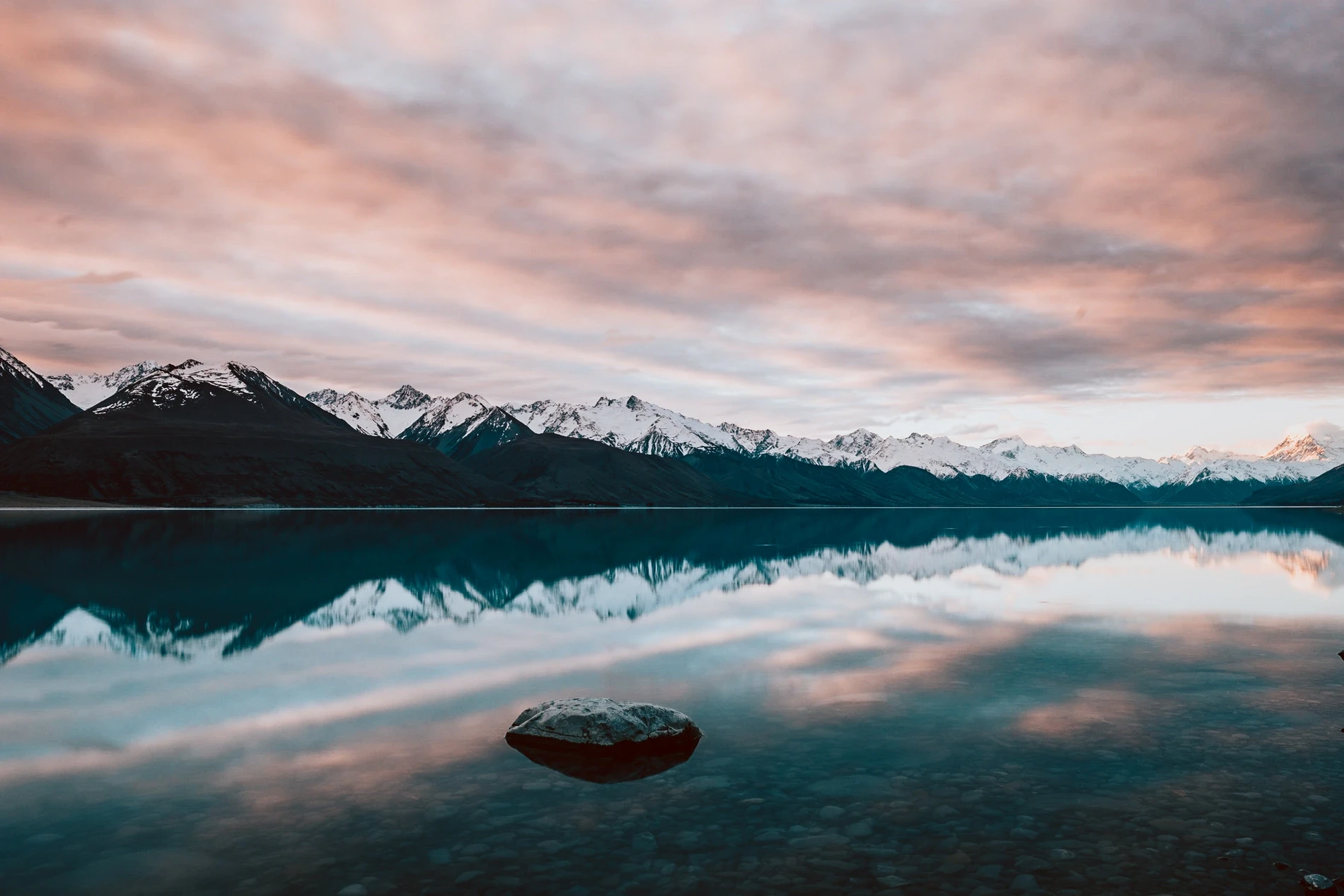 A small boat on a mirror-calm lake at dawn, pink cloud and snow-capped peaks reflected