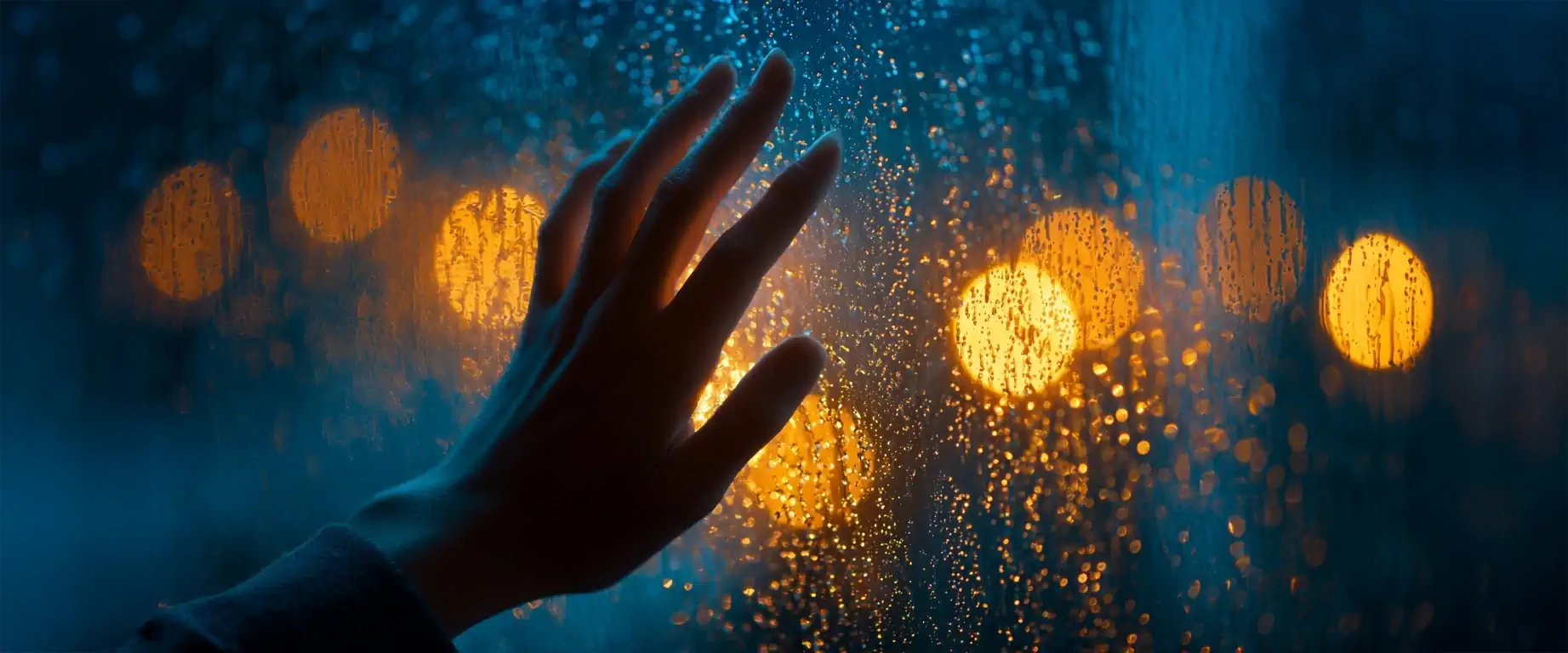 Main posée contre une fenêtre mouillée, avec des lumières floues en arrière-plan, symbolisant la recherche de présence intérieure.