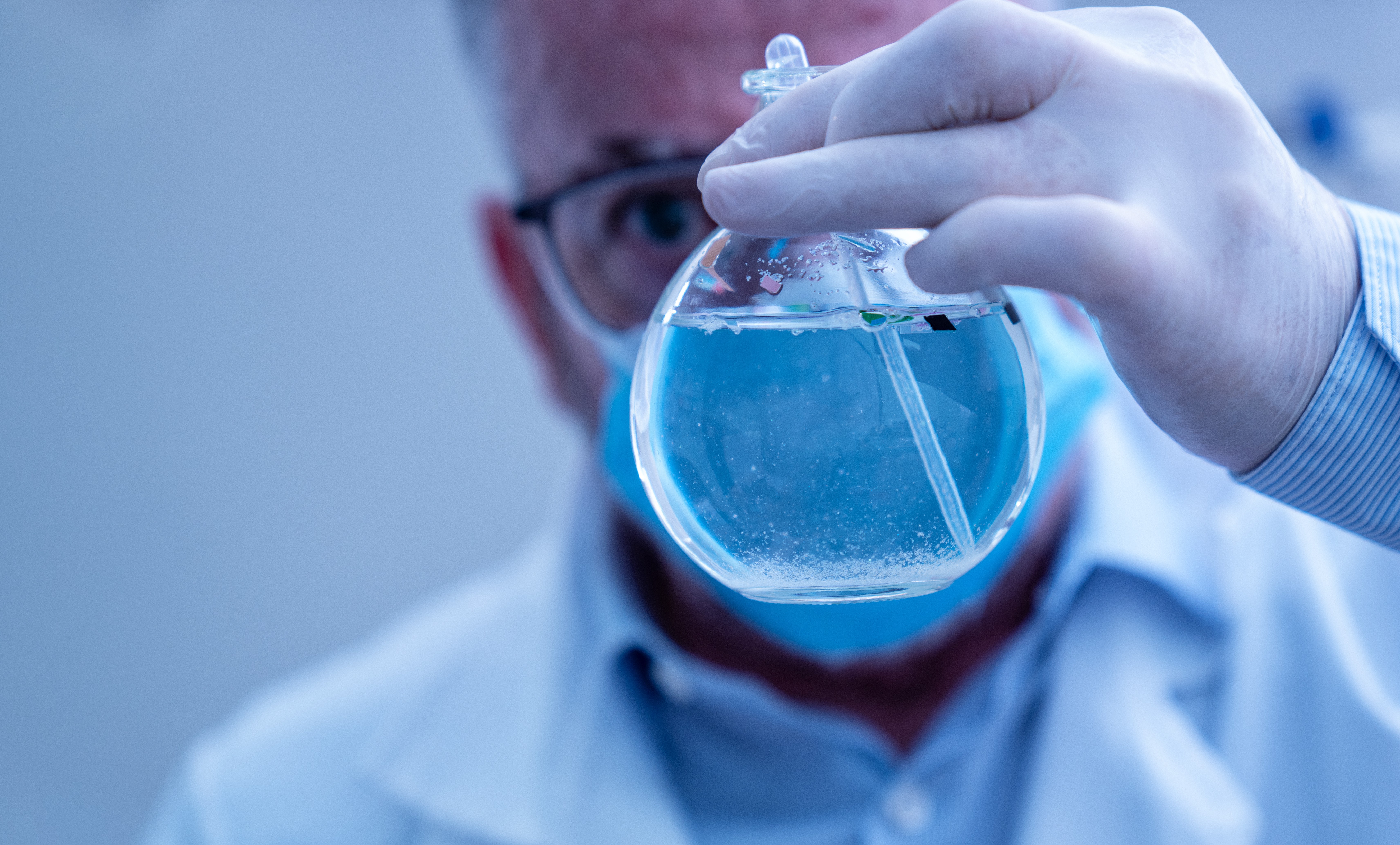 Scientist wearing mask and gloves holding a flask containing blue liquid.