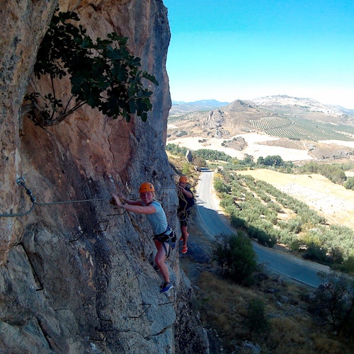 两名佩戴头盔和安全带的人攀登陡峭的岩壁,背景是风景如画的乡村景观。