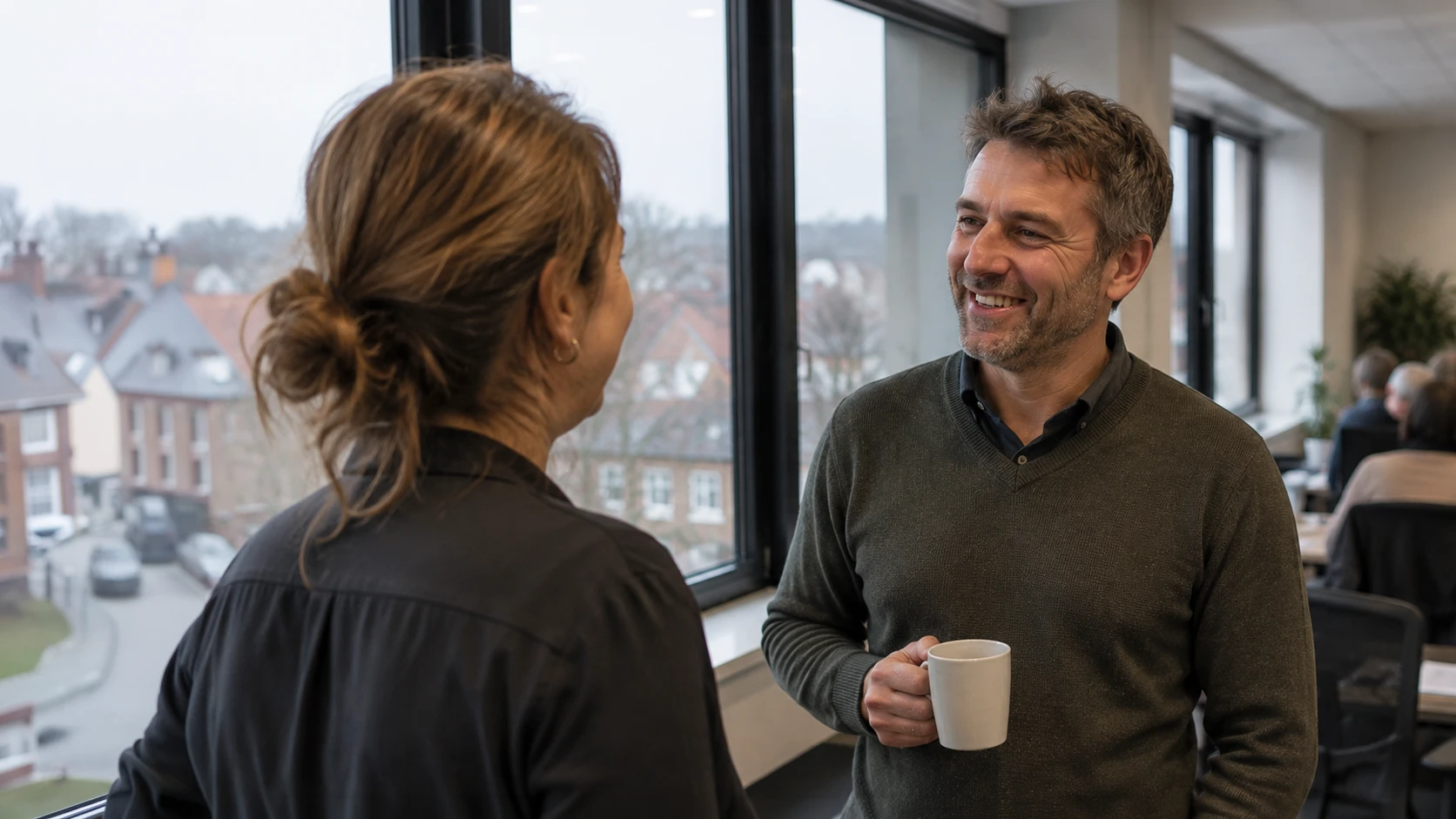 Two colleagues chatting candidly in a Newcross office, one holding a coffee mug, with a team working in the background.