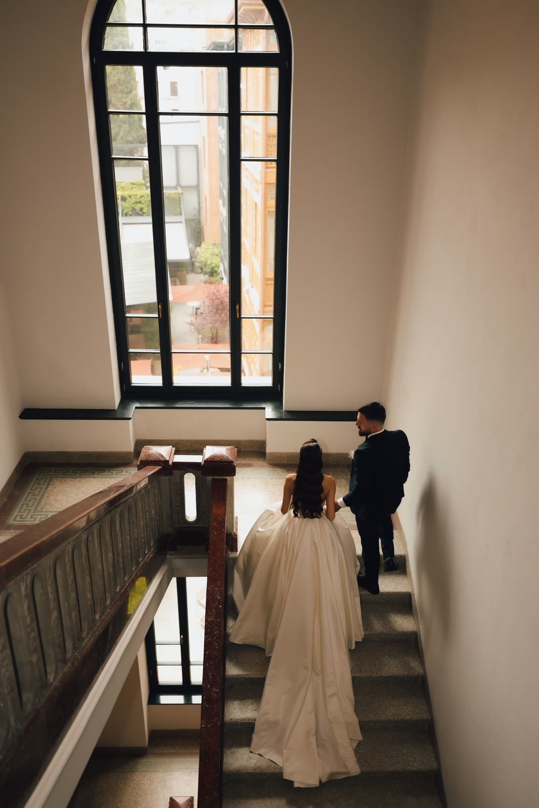 Bride and groom on a stair landing beside a tall window
