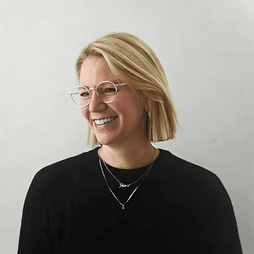 A person with wavy brown hair smiles against a light background, wearing a black blazer.