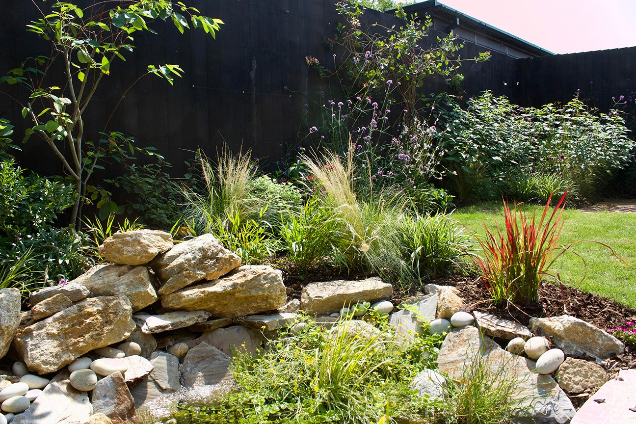 Lush garden with rocks, ornamental grasses, and greenery, featuring a dark building in the background.