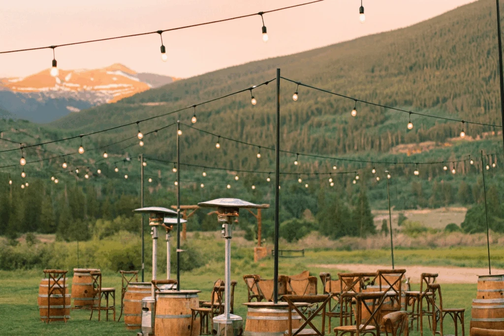 Rustic outdoor mountain wedding reception decorated with hanging string lights, wooden barrel tables, and patio heaters, overlooking sweeping alpine views and a golden sunset.