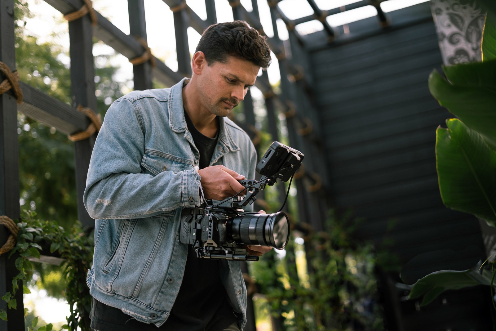 A person in a light jacket stands in a greenhouse, focused on examining a plant with a tool.