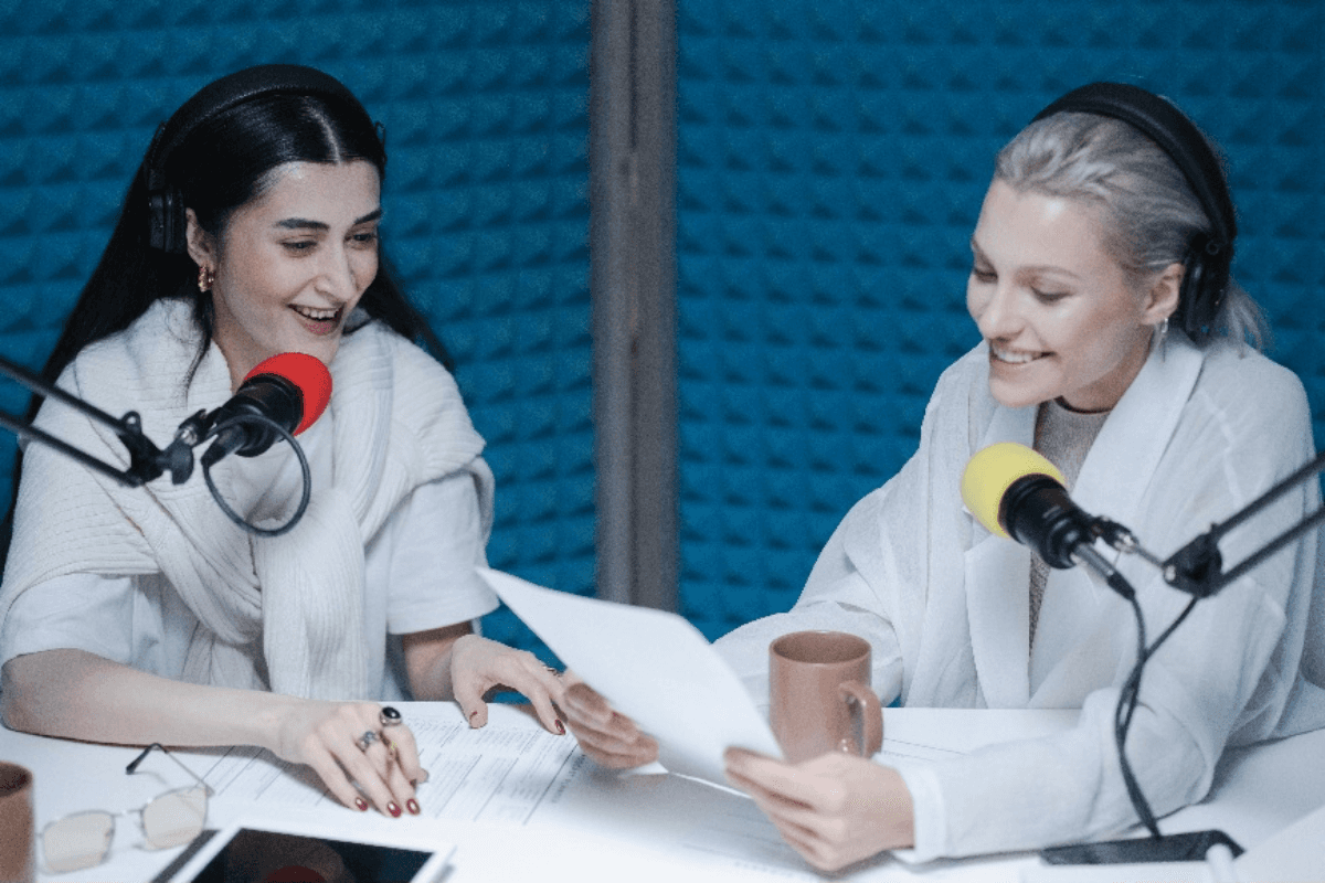 Two women in a recording studio with microphones, laughing and reading from a paper during a podcast session.