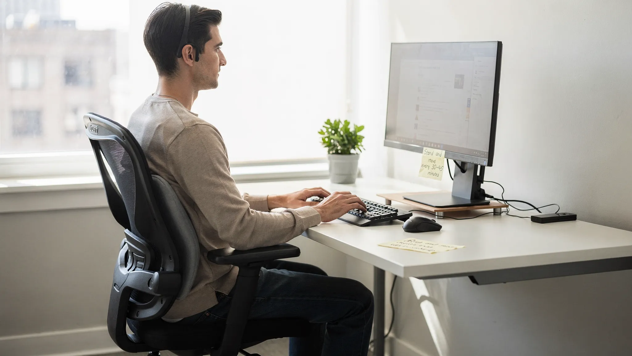 A person working at a desk with neutral seated posture, feet flat, hips slightly above knees, lumbar support, and a reminder note to stand and move every 30-45 minutes.