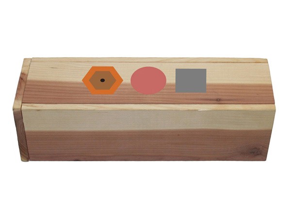 A rectangular wooden box with light-colored horizontal bands and darker reddish-brown horizontal bands. Three colored shapes are placed on the box: an orange hexagon with a black dot, a red circle, and a gray square. The box is positioned horizontally against a plain white background.