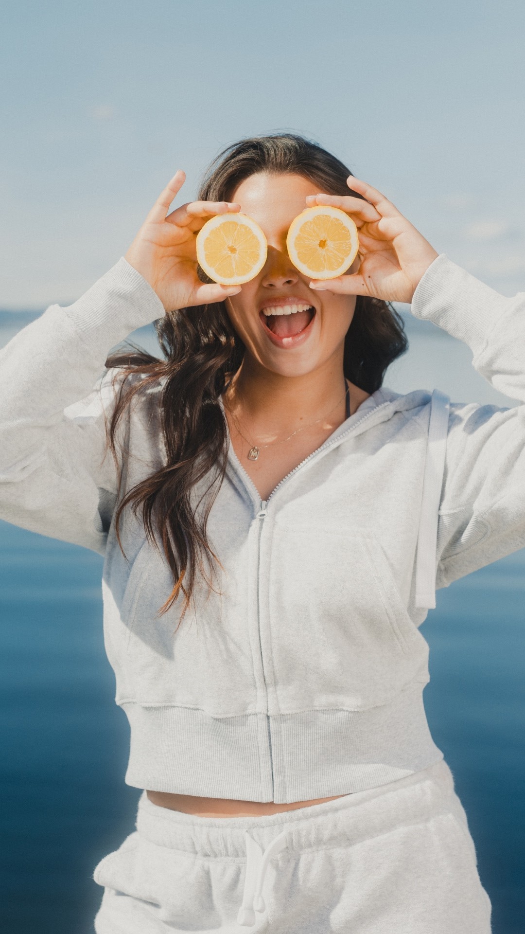 Young woman dressed in light grey ziphood, holding two lemons in front of her eyes and smiling