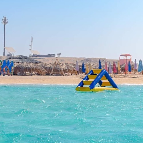 Inflatable water structure floating in turquoise sea with a sandy beach and thatched umbrellas in the background.