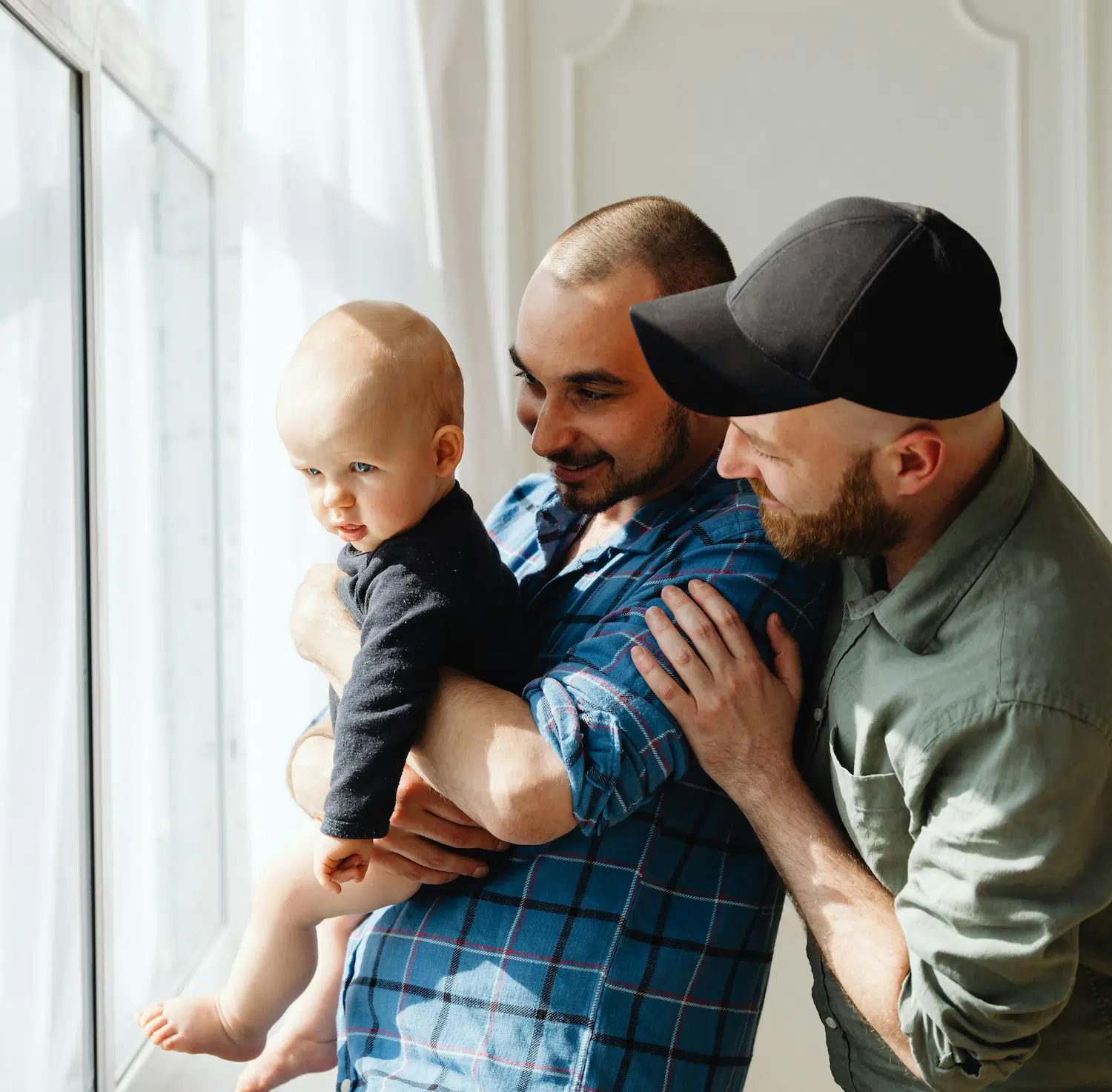 Two fathers holding their baby by a window, symbolizing LGBTQ+ family therapy and affirming counseling in NYC