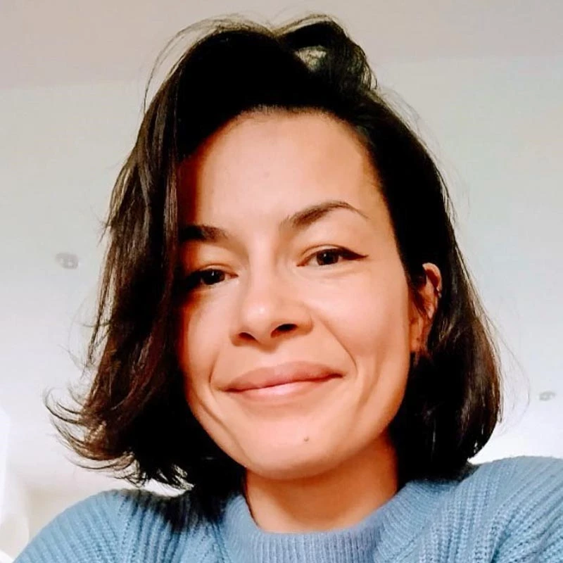 A person with short dark hair wears a light blue knit sweater. The background shows an indoor setting with soft, neutral lighting.