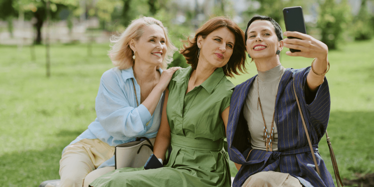 Three women in their 40s and 50s sitting outdoors, smiling, and taking a selfie together in a relaxed, empowered moment.