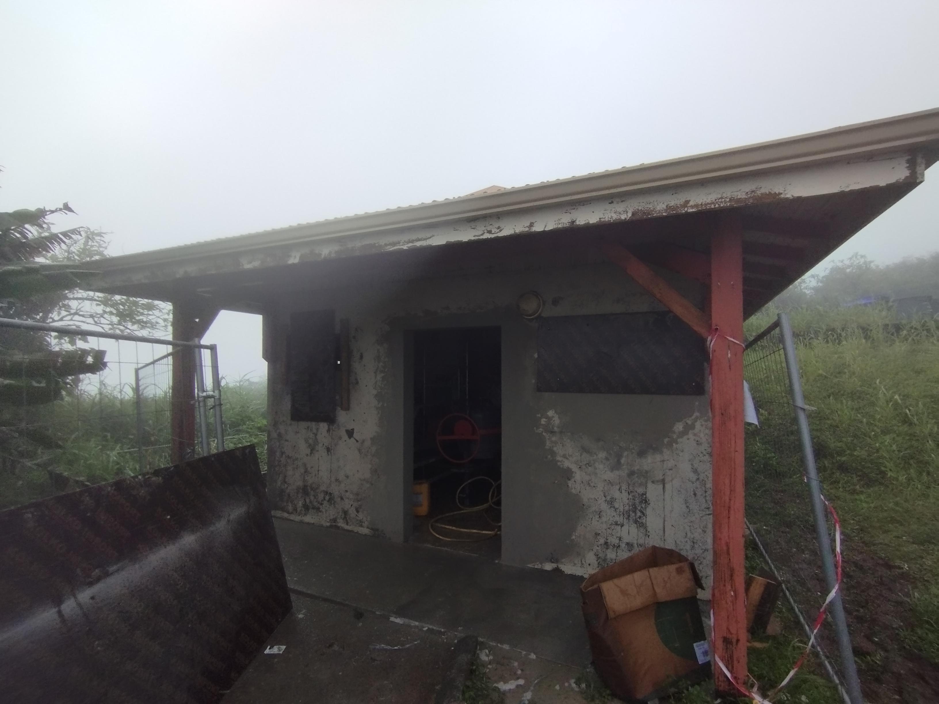 Petite maison en béton usé par les intempéries avec un toit rouillé dans un environnement brumeux. L'herbe envahissante qui l'entoure crée une atmosphère de désolation et d'abandon.
