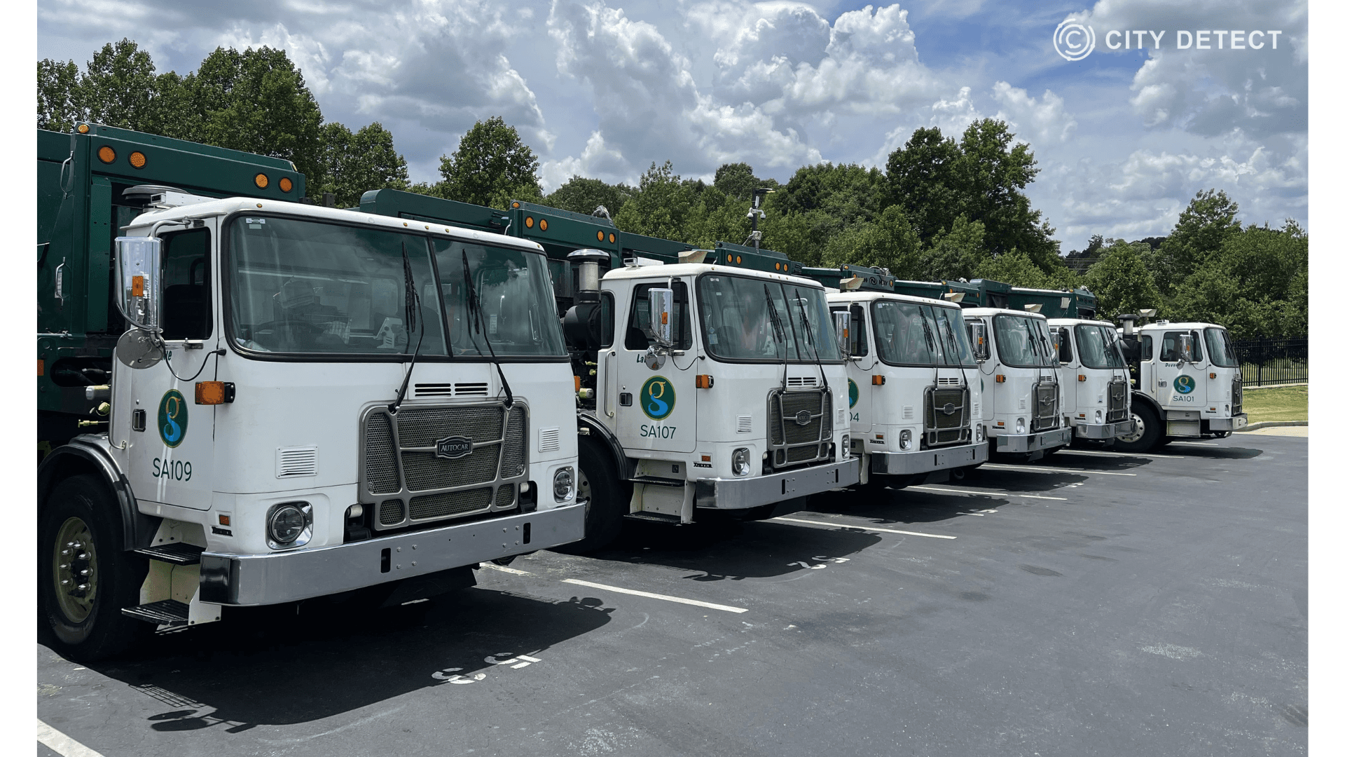 A line of white City Detect garbage trucks are parked in a row on a sunny day with trees in the background.