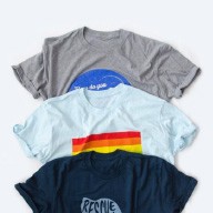 Three t-shirts stacked on top of each other. The top shirt is gray, the middle is light blue, and the bottom is navy. Each shirt has a different design.