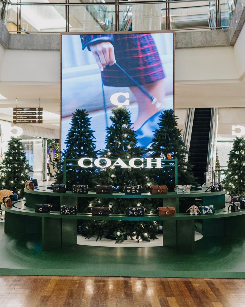 A festive display featuring a green circular showcase with various handbags by Coach, surrounded by Christmas trees, in a shopping mall setting.