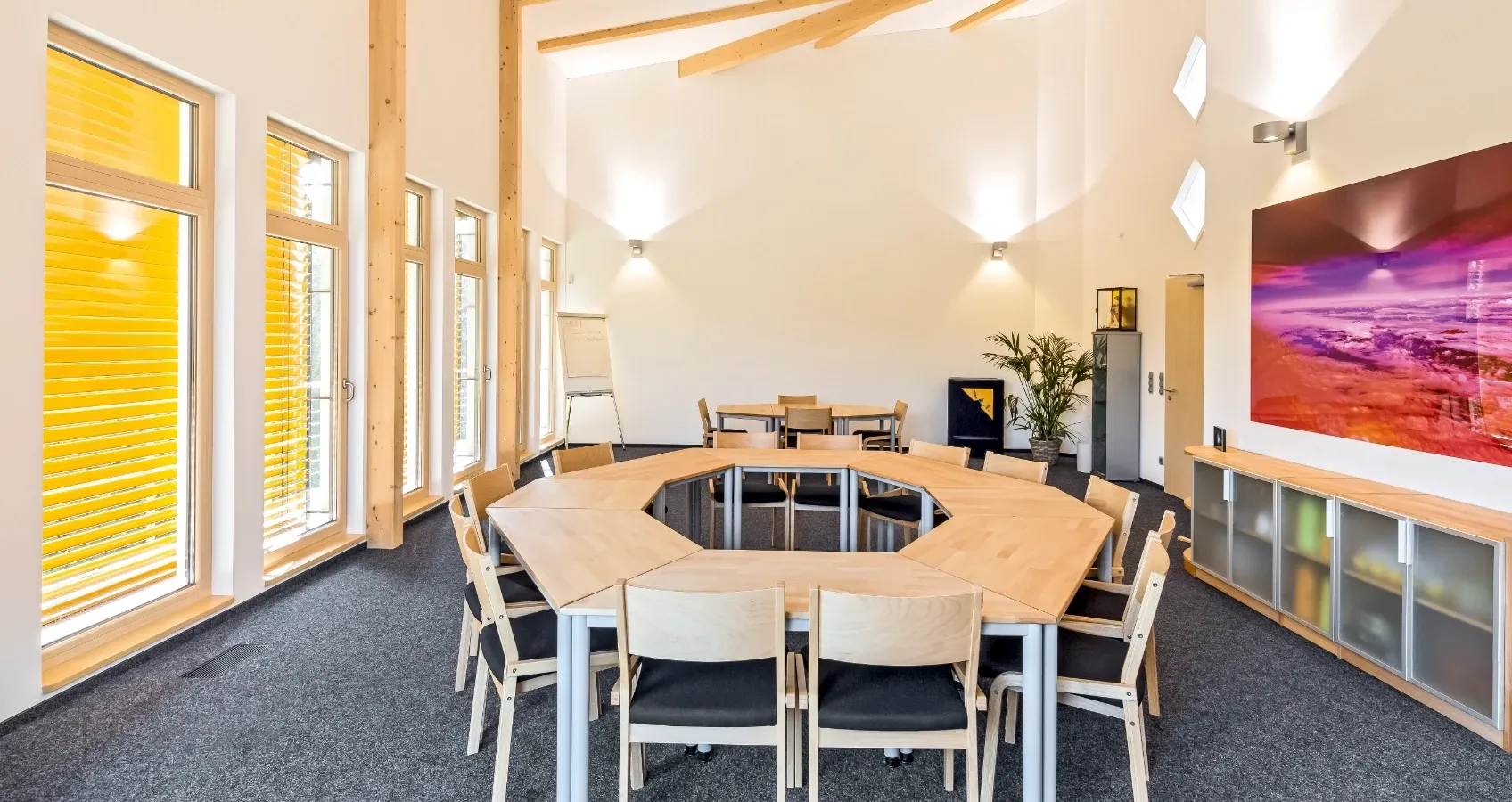Konferenzraum mit oktogonalem Tisch und Stühlen, helle Wände, Holzdeckenbalken und große Fenster mit gelben Jalousien.