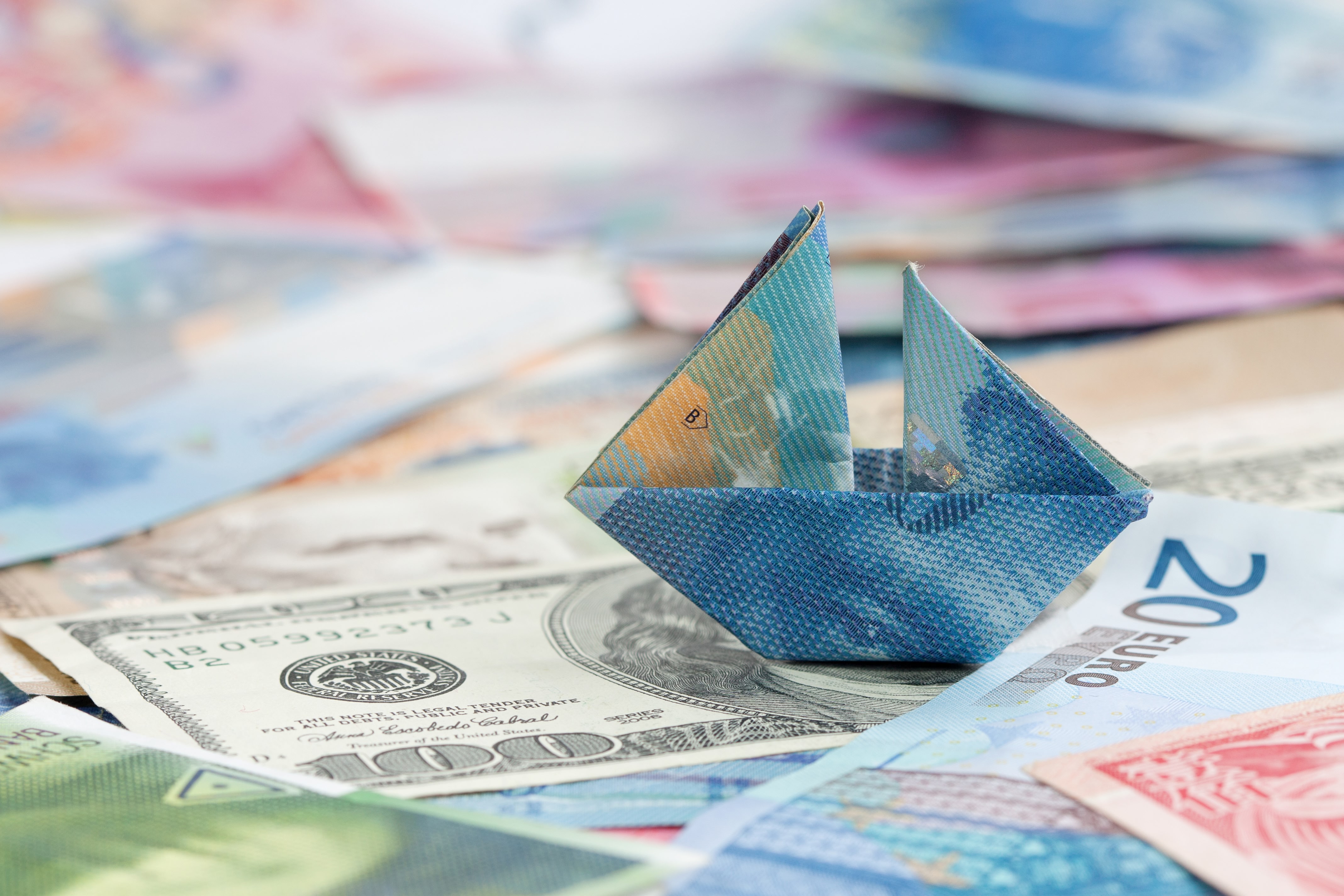 Un billet de 100 CHF plié en forme de bateau en papier, posé sur des billets de banque étrangers.