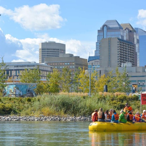 A group of people in life vests are on a yellow raft in a river, with a cityscape and greenery in the background.